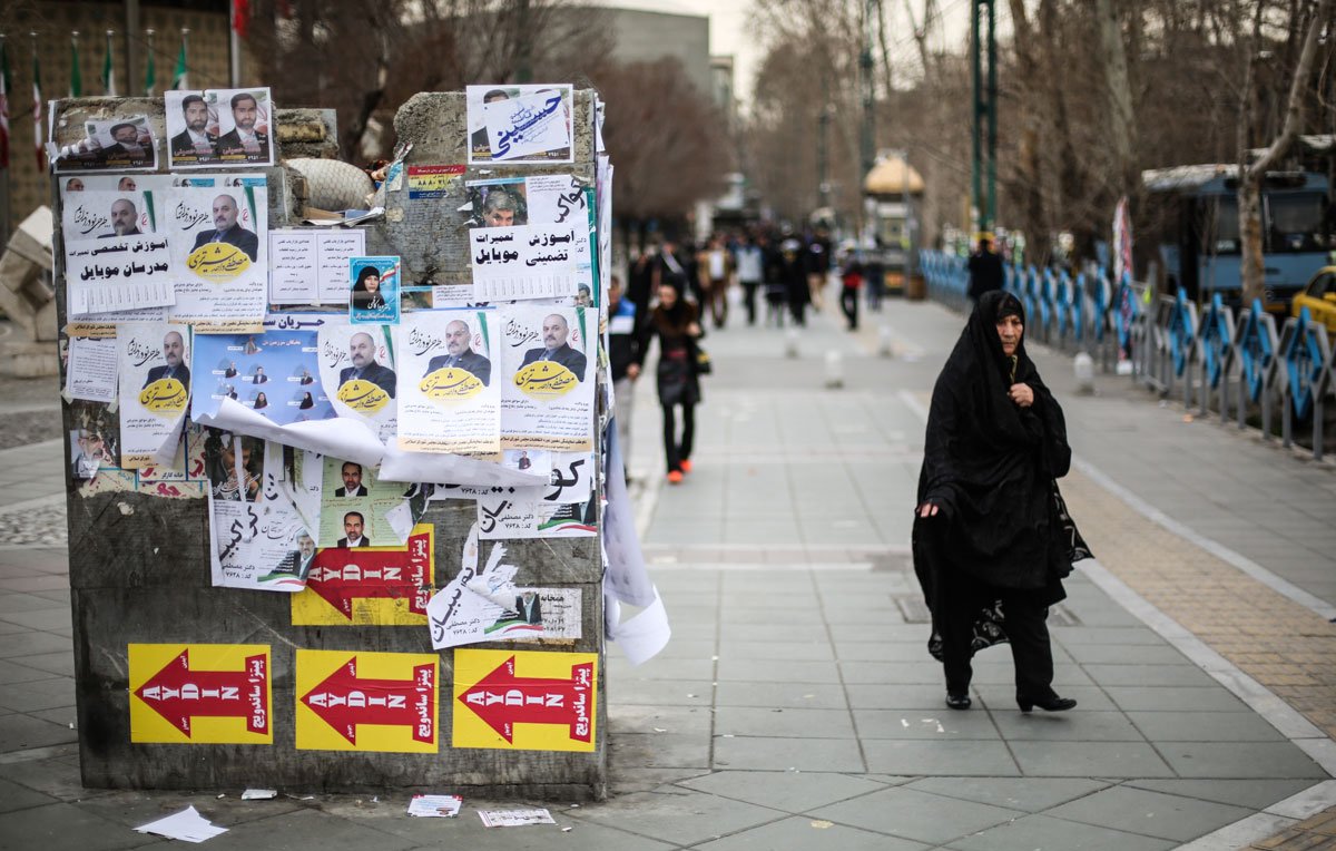 L'image montre une rue animée où une femme portant un hijab noir marche sur le trottoir. À côté d'elle, un mur est recouvert d'affiches et de banderoles, certaines de couleur vive, d'autres jaunies et déchirées. Les affiches semblent inclure des images de personnes et des slogans en écriture arabe. En arrière-plan, on peut distinguer des bus et des passants, créant une atmosphère urbaine. Le sol est en pavé, et les arbres dépouillés suggèrent une saison froide.