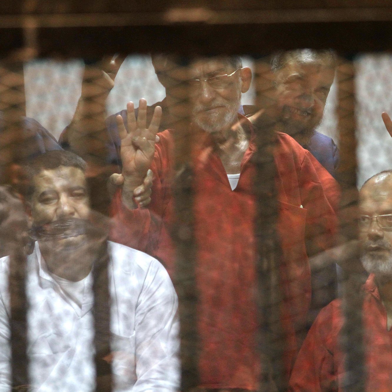 L'image montre un groupe d'hommes derrière une grille ou un panneau de verre. Ils semblent être dans un contexte de détention ou de procès, avec des expressions variées sur leurs visages. Certains lèvent les mains, suggérant un geste de protestation ou de solidarité. L'ambiance générale est sombre, évoquant des thèmes de justice et de droits humains.