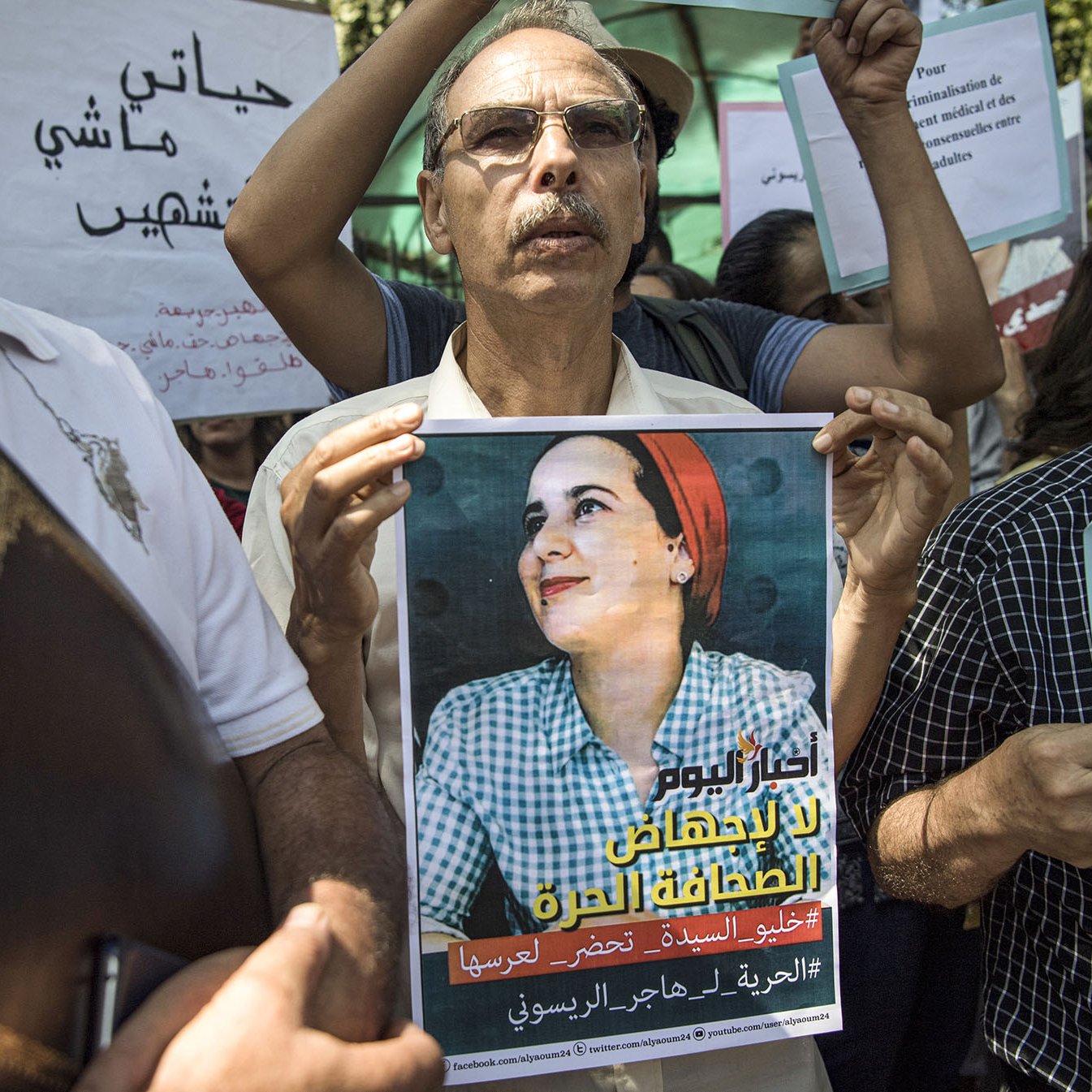L'image montre un groupe de personnes rassemblées pour une manifestation. Au premier plan, un homme tient une affiche qui représente une femme avec un sourire, probablement une figure symbolique pour le mouvement. Les participants, majoritairement des adultes, tiennent également des pancartes avec des slogans en arabe et en français, indiquant leur lutte ou leur soutien à une cause, sans doute liée à des droits ou à une injustice. L'atmosphère semble déterminée et engagée.