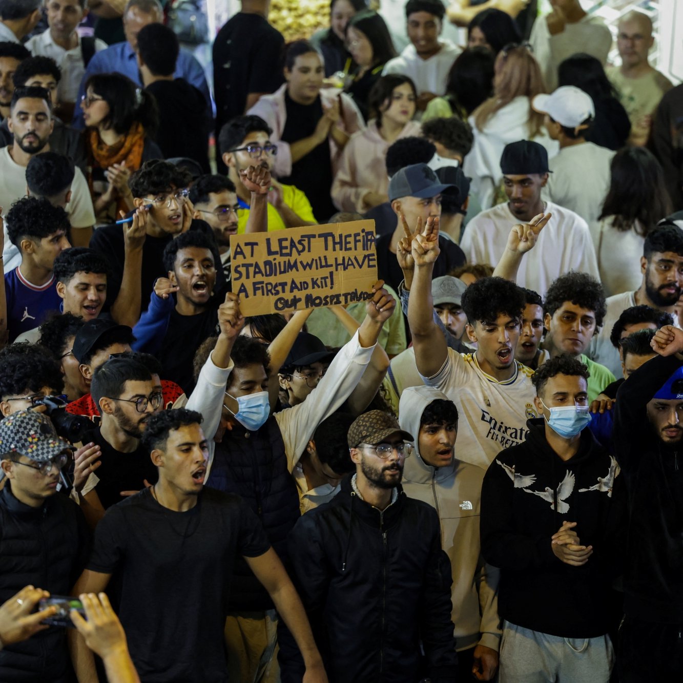 حشود من الناس في مكان مزدحم، يرفعون لافتة ويهتفون، مع تباين في الألوان والملابس.