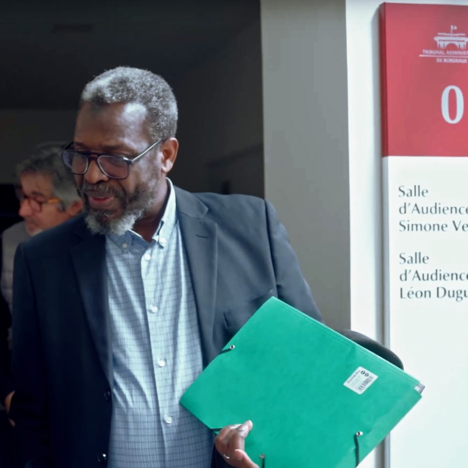 L'image montre un groupe de personnes dans un couloir apparemment nimbé d'une ambiance professionnelle. Deux hommes au premier plan interagissent, l'un tenant un téléphone et l'autre un dossier vert. L'environnement semble être celui d'un bâtiment public ou d'un tribunal, avec des panneaux indiquant les salles d'audience. Les visages des personnes sont engagés dans une conversation, ce qui suggère un échange collaboratif ou une discussion importante.