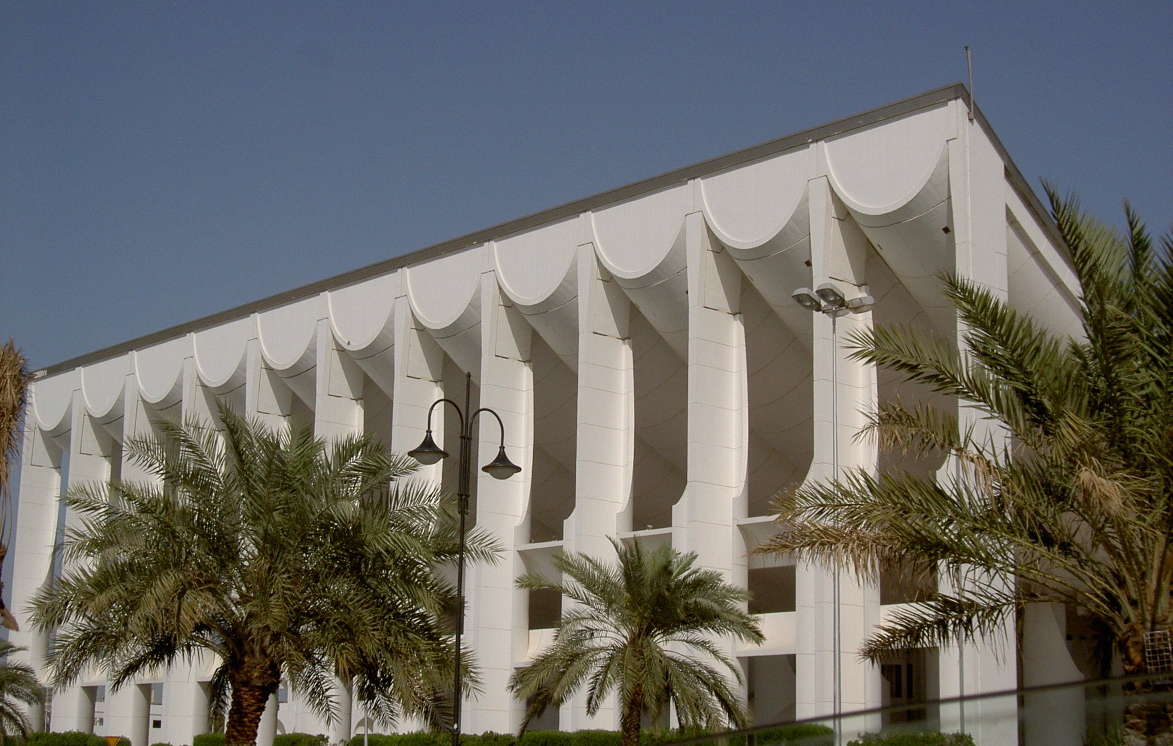 الصورة تظهر مبنى معماري متميز يتميز بتصميمه العصري. يتكون المبنى من واجهة بيضاء تحتوي على عموديات بارزة، مع استخدام الأقمشة البيضاء التي تزين السقف وتضفي عليه مظهراً فريداً. تحيط بالمبنى أشجار النخيل والحدائق الخضراء، مما يضفي جوًا منعشًا وجميلًا. السماء زرقاء صافية مما يعكس جمال اليوم.