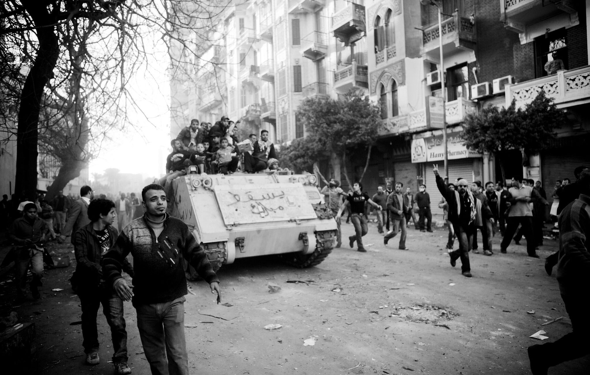L'image montre une scène de manifestation urbaine, avec des gens dans une rue animée. Au premier plan, plusieurs manifestants marchent, certains levant les bras en l'air. À l'arrière-plan, un tank est visible, portant des inscriptions. La scène est en noir et blanc, ce qui intensifie l'atmosphère dramatique et chargée d'émotion. Les bâtiments environnants et les arbres ajoutent à l'environnement urbain. L'ensemble dégage un sentiment de tension et de mouvement.