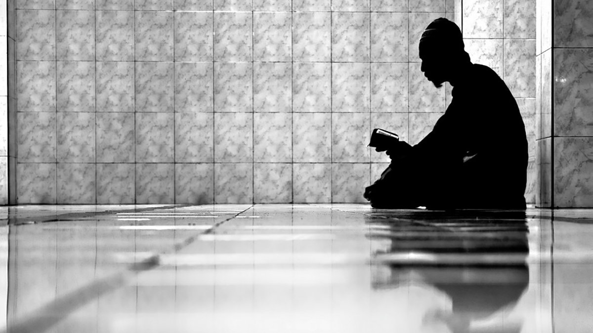 L'image montre un individu assis sur le sol, plongé dans la lecture d'un livre. La composition est en noir et blanc, créant un contraste fort entre l'ombre de la personne et l'éclat des carreaux brillants au sol. L'ambiance est calme et contemplative, et le reflet de la silhouette sur le sol ajoute une dimension introspective à la scène. Les murs en arrière-plan sont recouverts de carrelage, ce qui renforce l'aspect minimaliste de l'image.