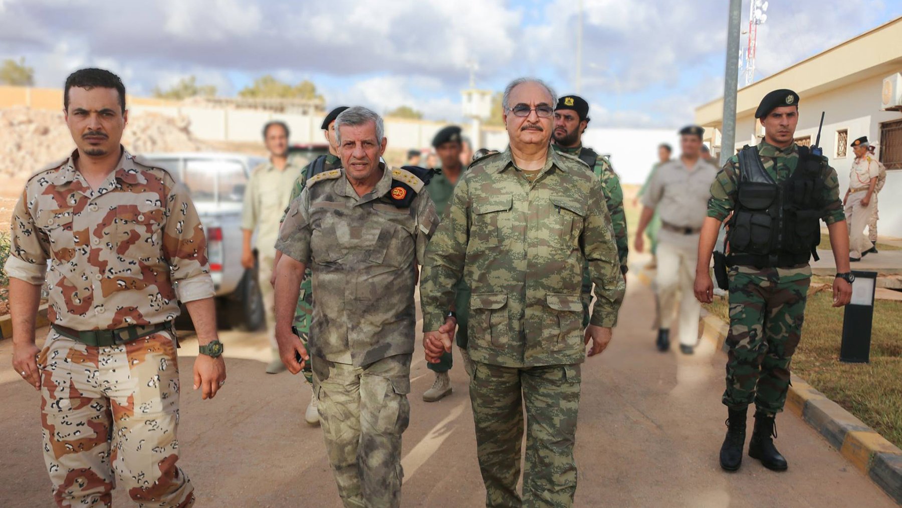 L'image montre un groupe de militaires marchant ensemble dans un environnement urbain. Deux hommes en avant-plan semblent discuter tout en marchant côte à côte. L'un d'eux porte un uniforme camouflage de couleur verte, tandis que l'autre a un uniforme militaire gris avec des distinctions. En arrière-plan, on peut voir d'autres soldats et des véhicules militaires, ce qui indique qu'ils se trouvent dans une zone potentiellement sécurisée. Les soldats semblent concentrés et en mission.