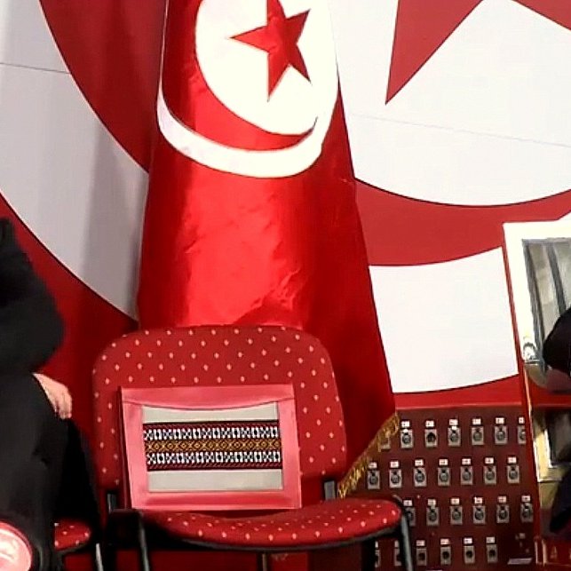 L'image montre deux hommes assis sur des chaises lors d'un événement. Ils semblent être en train d'écouter ou d'observer quelque chose, peut-être un discours ou une présentation. En arrière-plan, il y a un drapeau tunisien, ce qui indique que l'événement se déroule en Tunisie. L'un des hommes porte des lunettes et a une expression sérieuse, tandis que l'autre exerce une posture détendue. La scène semble officielle, peut-être liée à la politique ou à un rassemblement important.