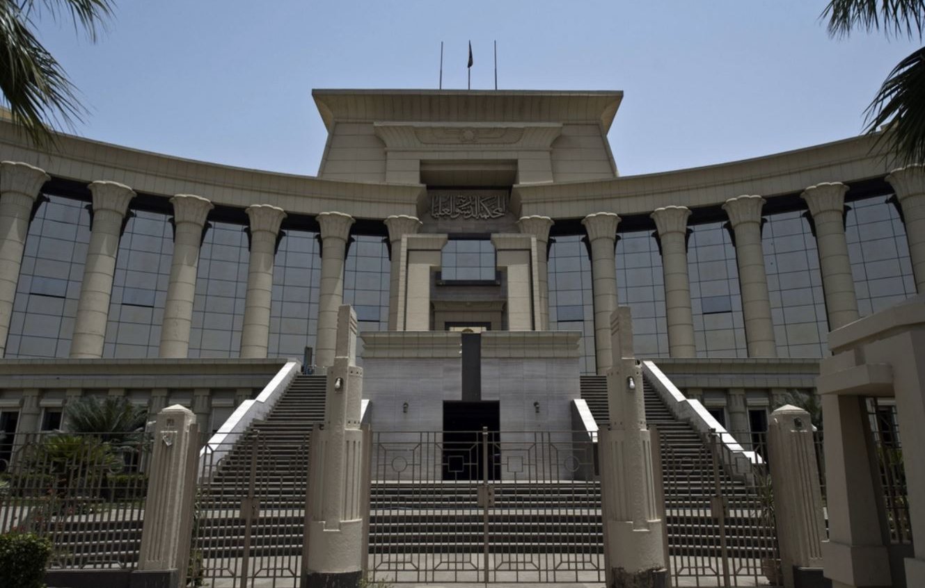 الصورة تظهر مبنى حكومي كبير يتميز بتصميم معماري حديث. يبرز في الصورة مدخل المبنى الذي يحتوي على مجموعة من السلالم والأعمدة الكبيرة. الواجهة تمتاز بالنوافذ الزجاجية العريضة التي تعكس الضوء، وتوجد أعلام مرفوعة فوق السطح. المنطقة المحيطة بالمبنى تحتوي على أشجار ونباتات تعطي جواً من الخضرة. يبدو الطقس مشمساً وواضحاً، مما يضفي على المشهد حيوية.