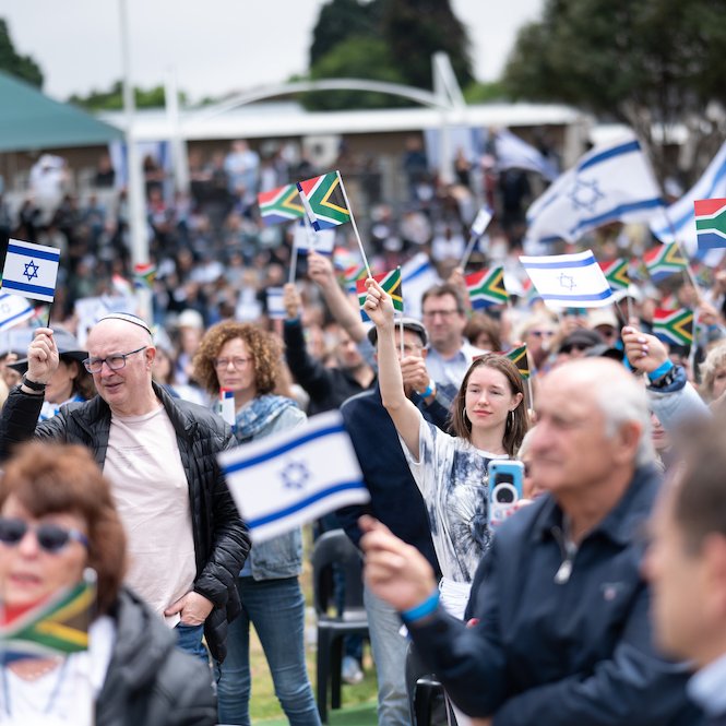 L'image montre une grande foule rassemblée, tenant des drapeaux israéliens et sud-africains. Les personnes semblent engagées et heureux, certaines souriant et levant les drapeaux en signe de soutien. L'environnement est un espace extérieur, et on peut voir des stands ou des tribunes en arrière-plan, suggérant un événement public ou une cérémonie. L'atmosphère semble festive et solidaire.