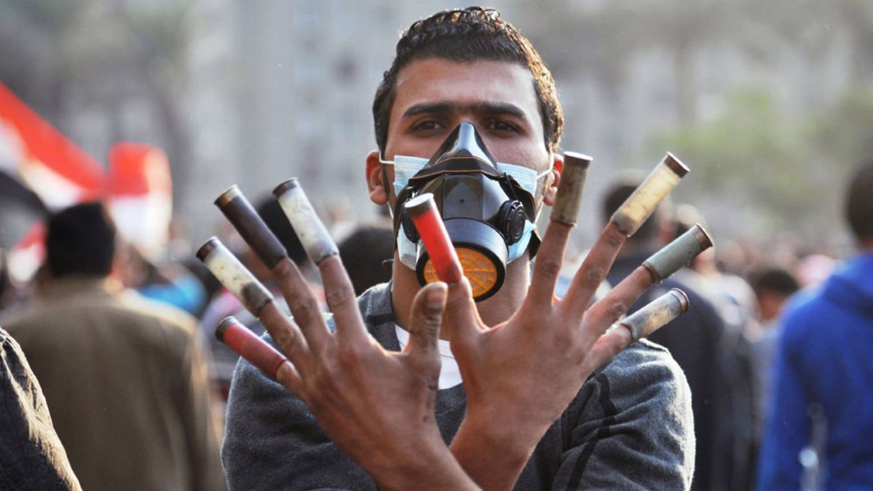L'image montre un homme portant un masque de protection, levant les mains avec plusieurs cartouches de gaz lacrymogène visibles. Son expression semble déterminée, et l'arrière-plan indique qu'il est présent dans une foule, probablement lors d'une manifestation. L'atmosphère de la scène semble être celle d'un événement intense et chargé d'émotion.