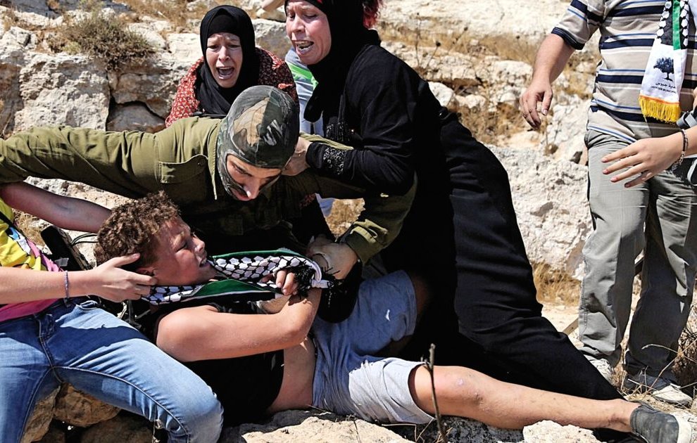 تصویر نشان‌دهنده یک صحنه تنش‌زا است که در آن چند نفر در حال مقاومت یا کمک به یکدیگر هستند. تعدادی از افراد در حال تعامل با یک نظامی به نظر می‌رسند و برخی از آنها به وضوح نگران و هیجان‌زده به نظر می‌رسند. این تصویر ممکن است به یک واقعه یا اعتراض خاص مرتبط باشد. زمینه صحنه طبیعی و سنگی است که احساس وخامت موقعیت را بیشتر می‌کند.