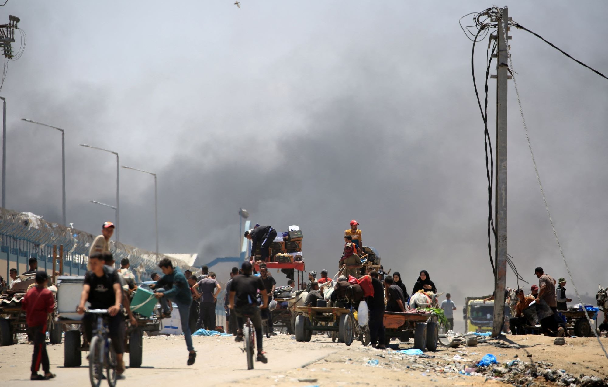 L'immagine mostra una scena di un'area urbana colpita da un conflitto o una crisi. Si vedono diverse persone che si muovono lungo una strada, alcune in bicicletta, altre a bordo di carri trainati da animali. Sullo sfondo, si nota del fumo denso che si alza, suggerendo un incendio o una situazione pericolosa. Le persone sembrano essere coinvolte in attività di trasporto, mentre l'atmosfera è tesa e caotica, con una presenza significativa di detriti e rifiuti sulla strada.