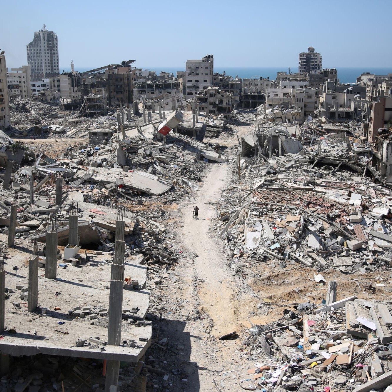 L'image montre une scène de destruction dans une zone urbaine après un conflit. De nombreux bâtiments sont partiellement détruits, avec des débris éparpillés sur le sol. On peut apercevoir des ruines de constructions, tandis qu'une route poussiéreuse traverse le paysage. À l'horizon, on voit la mer, ce qui contraste avec la gravité des destructions. L'atmosphère apparaît calme, mais la destruction est évidente et témoigne des effets dévastateurs de la guerre.