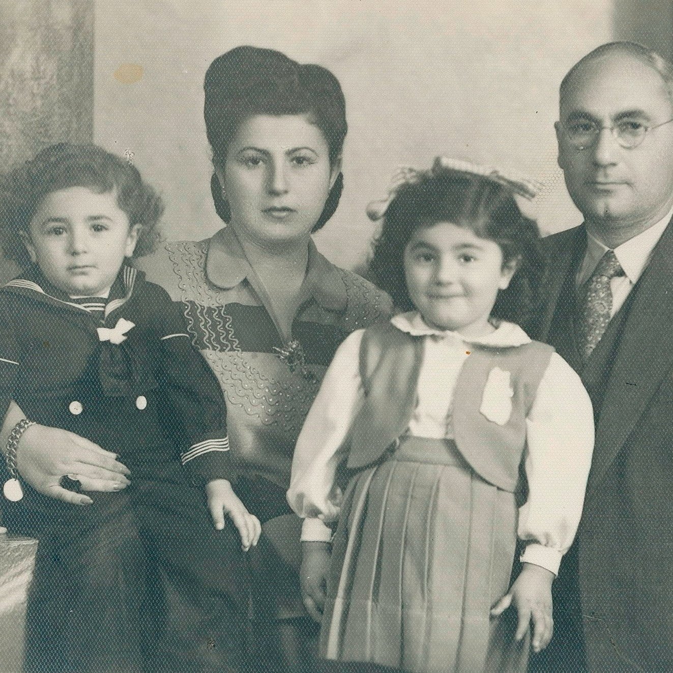 تظهر في الصورة عائلة مكونة من أربعة أفراد. الأم ترتدي زيًا تقليديًا وتبدو أنيقة، بينما الأب مرتديًا بدلة رسمية ونظارات. يوجد طفلان، ولد فتاة، يقفان أمام والديهما. يُظهر تعبير الوجه الودود والمشاعر الأسرية الدافئة. الخلفية بسيطة وتضيف لمسة كلاسيكية للصورة، التي تبدو قديمة.