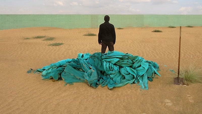 L'image montre un paysage désertique avec des dunes de sable sous un ciel partiellement nuageux. Au premier plan, un homme se tient dos à la caméra sur un monticule de tissus turquoise. En arrière-plan, on aperçoit un mur vert qui contraste avec le sable. L'ensemble de la scène évoque une atmosphère d'isolement et de contemplation.
