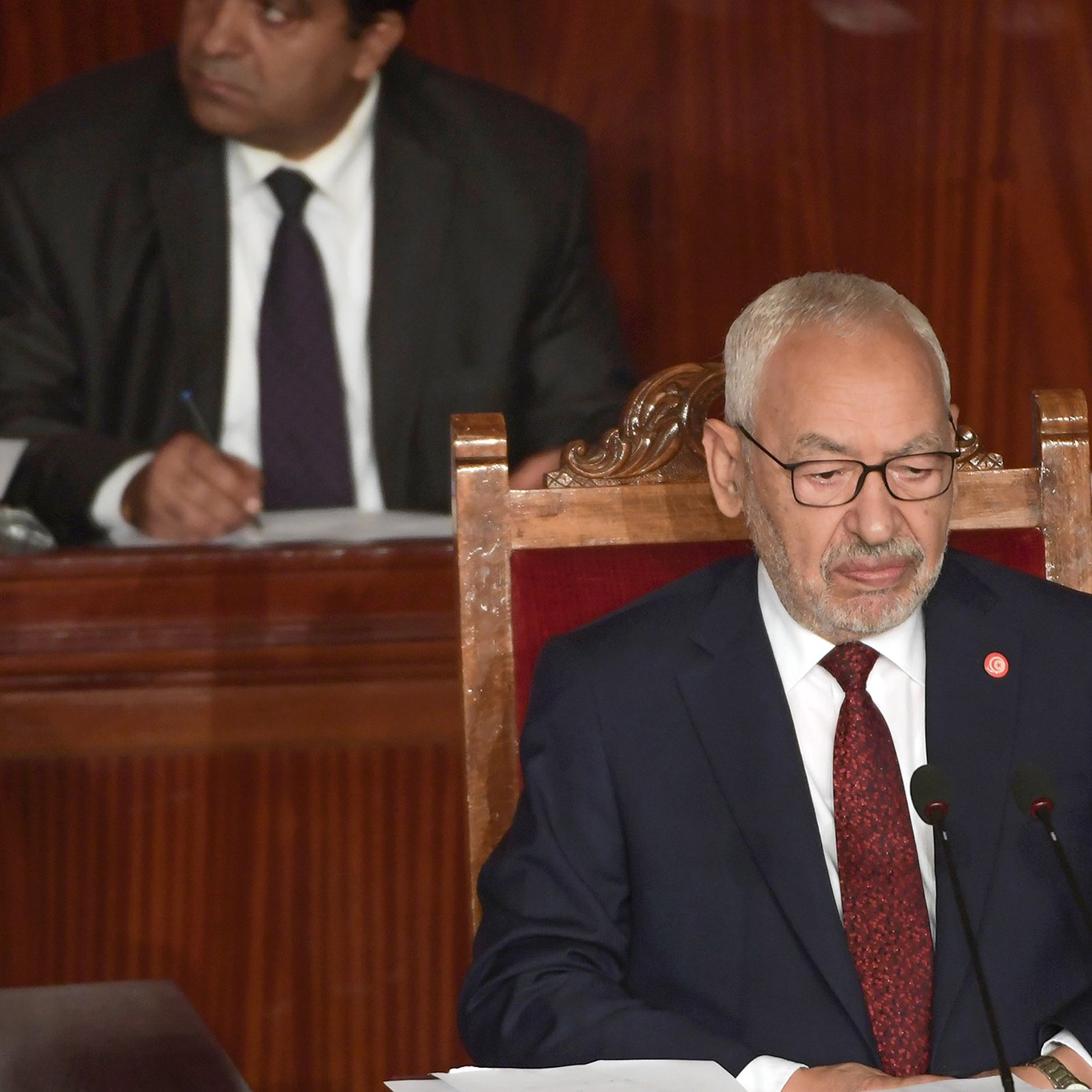 الصورة تظهر رجلًا يرتدي بدلة رسمية مع نظارات، يجلس على كرسي أمام منصة. يبدو أنه في جلسة رسمية أو برلمانية، حيث توجد ميكروفونات أمامه. في الخلفية، يظهر رجل آخر يرتدي بدلة أيضًا، يبدو أنه يستمع أو يتابع ما يحدث. الخلفية تتميز بتفاصيل خشبية، مما يضفي طابعًا رسميًا على المكان.