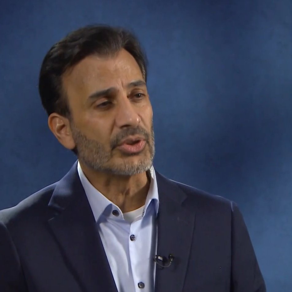 Nell'immagine si vede un uomo con una barba curata e capelli scuri, vestito con una giacca scura e una camicia chiara. Sembra che stia partecipando a un'intervista o a un discorso, mentre si esprime con attenzione. Sullo sfondo c'è un'ambientazione di colore blu, che crea un'atmosfera professionale.