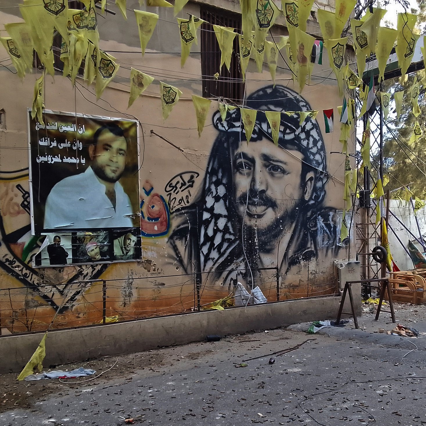 L'image montre un mur décoré d'une grande fresque représentant un homme avec un keffieh, typique de la culture arabe. À côté, il y a des affiches avec des portraits et des slogans. Des drapeaux jaunes pendent également, créant une ambiance festive ou militante. Le sol semble être en désordre avec divers objets autour, indiquant peut-être un événement ou une célébration récente. Les couleurs vives des décorations contrastent avec le mur beige.