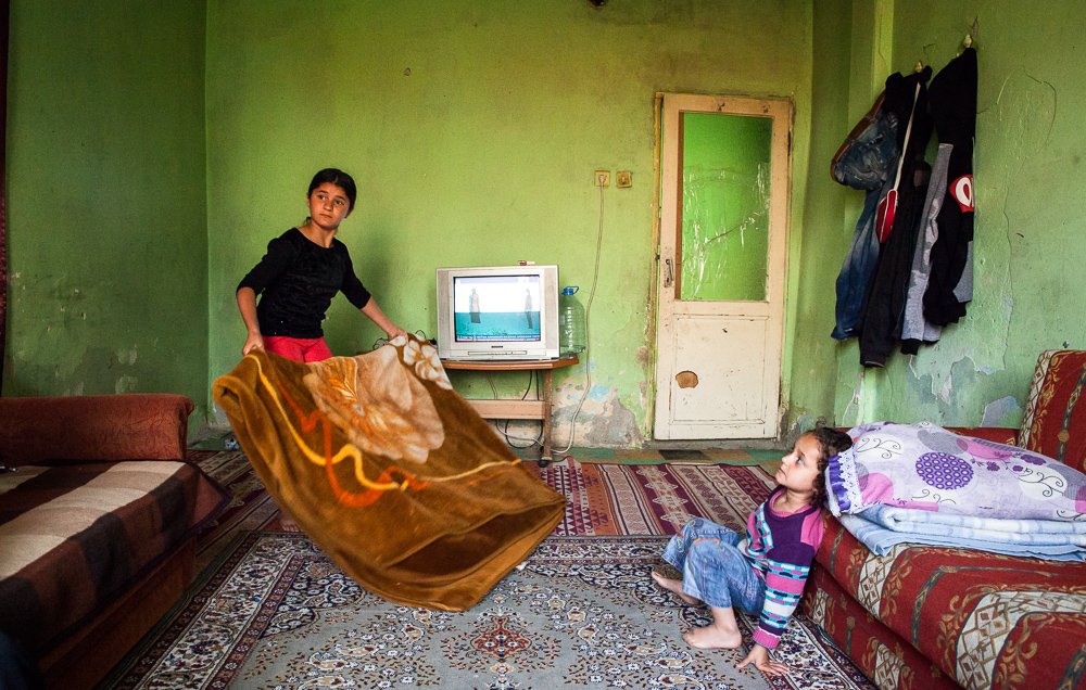 L'image montre une pièce modeste avec des murs verts. Au centre, deux enfants sont présents. L'un d'eux, un garçon, tient une grande couverture qu'il semble s'apprêter à lancer ou à étendre. L'autre enfant, une petite fille, est assise sur le sol, regardant le garçon avec curiosité. On peut voir un téléviseur allumé sur une petite table, et quelques vêtements accrochés au mur. Le décor est simple, avec un tapis et des canapés. L'ambiance générale semble familiale et décontractée.