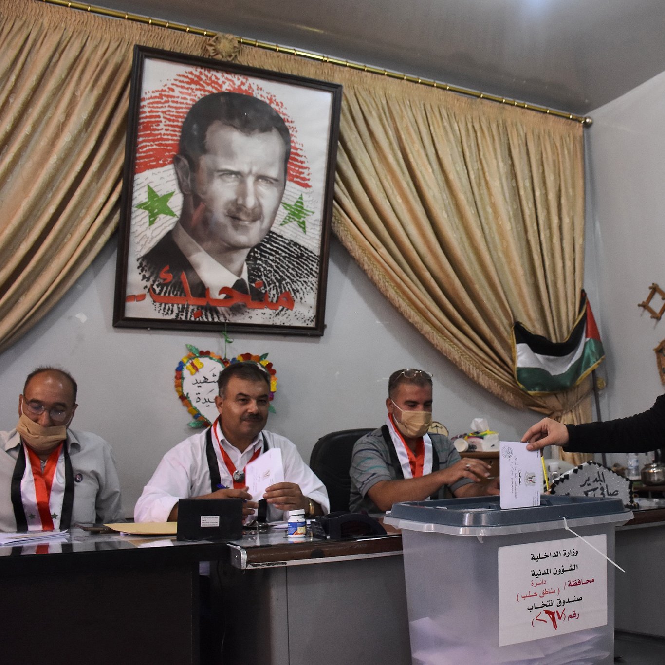 L'image montre une scène de vote, probablement lors d'élections en Syrie. Au premier plan, une femme en hijab dépose un bulletin dans une urne transparente. À l'arrière-plan, plusieurs hommes sont assis derrière une table, regardant le processus. Un grand portrait d'un homme, probablement un dirigeant, est accroché au mur, entouré de drapeaux et de décorations. Les participants portent des badges d'identification, et l'atmosphère semble sérieuse et formelle.