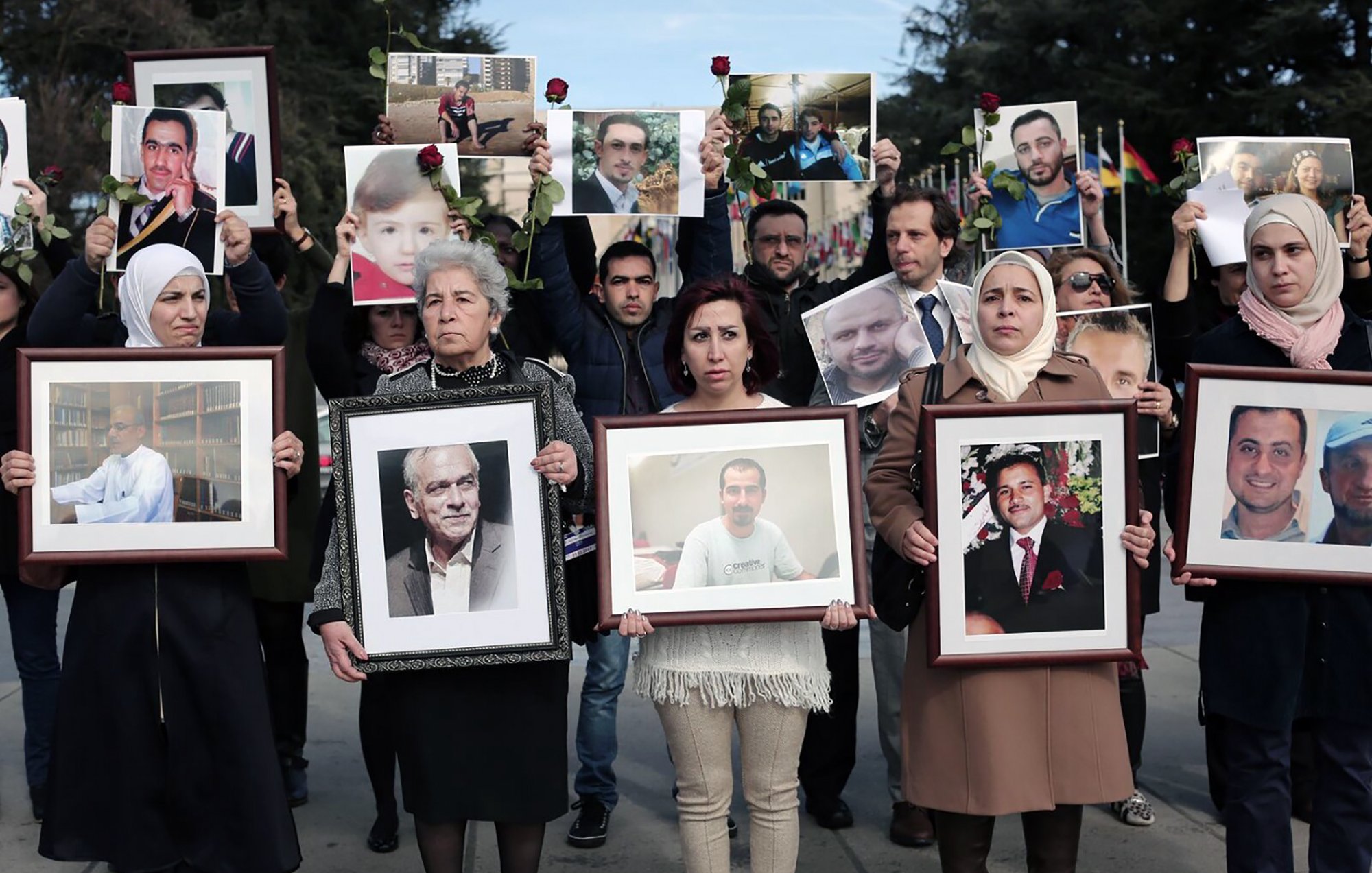 L'image montre un groupe de personnes participant à une manifestation ou un hommage. Elles tiennent des portraits de différents individus, certains avec des roses. Les expressions des participants semblent sérieuses et engagées, suggérant qu'ils rendent hommage à des personnes disparues ou aux victimes d'un conflit. Le décor en arrière-plan semble être en plein air, et plusieurs drapeaux sont visibles. Les vêtements des manifestants varient, avec quelques femmes portant des foulards.