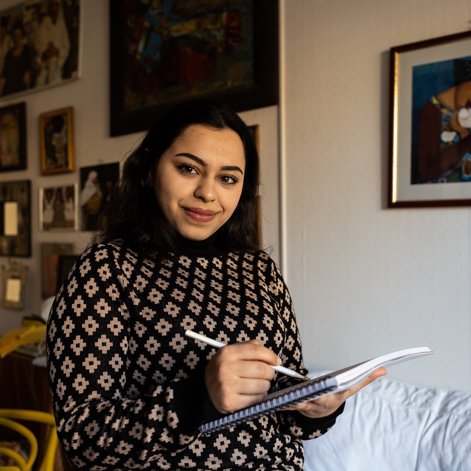 Nell'immagine, una giovane donna sorride mentre tiene un taccuino e una penna in mano. Indossa un maglione con un motivo geometrico. Sullo sfondo si può vedere una stanza con pareti decorate da vari quadri e fotografie. C'è un divano bianco e una sedia gialla, che aggiungono un tocco di colore all'ambiente. L'atmosfera è calda e accogliente.