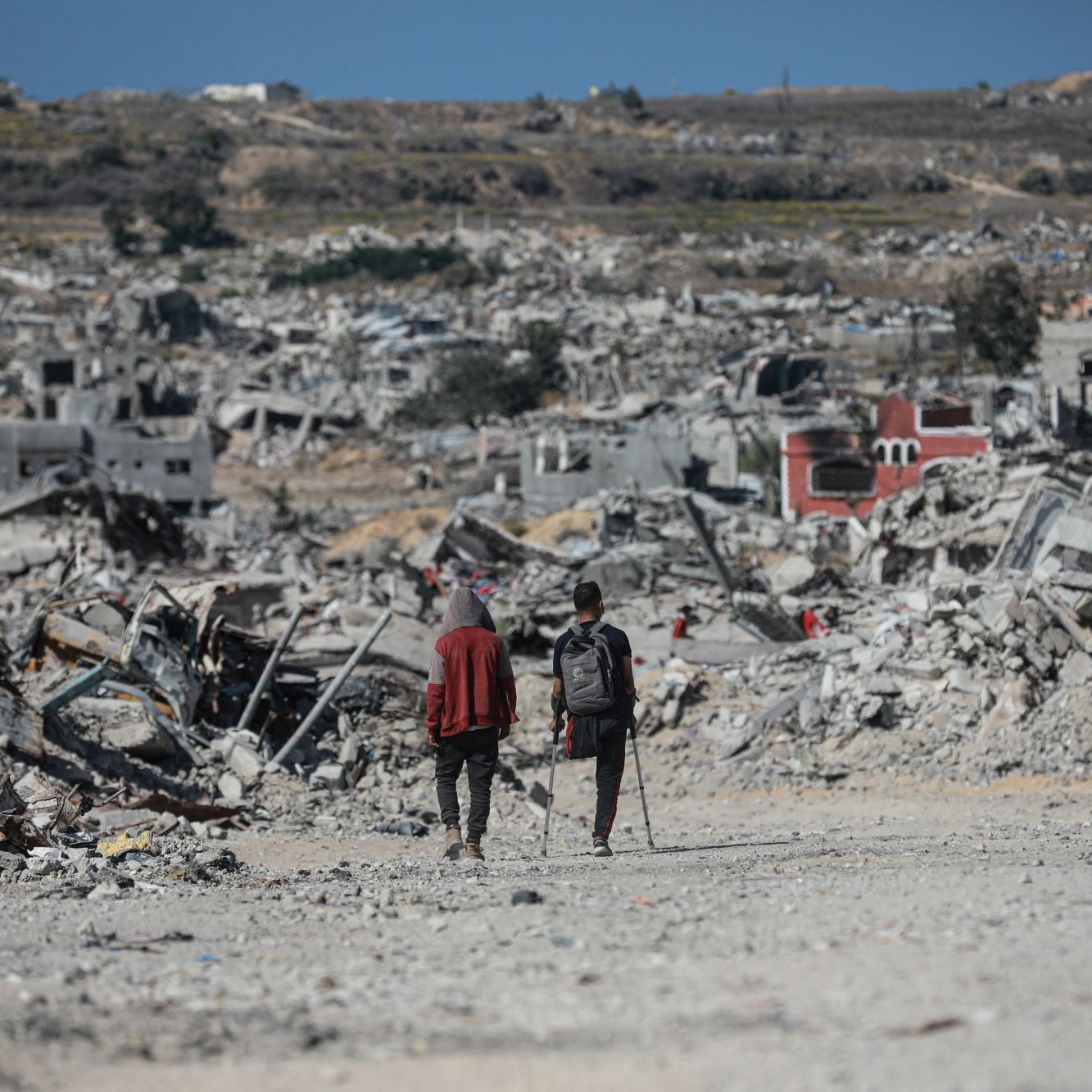 Due persone camminano tra le macerie di edifici distrutti in un paesaggio desolato.
