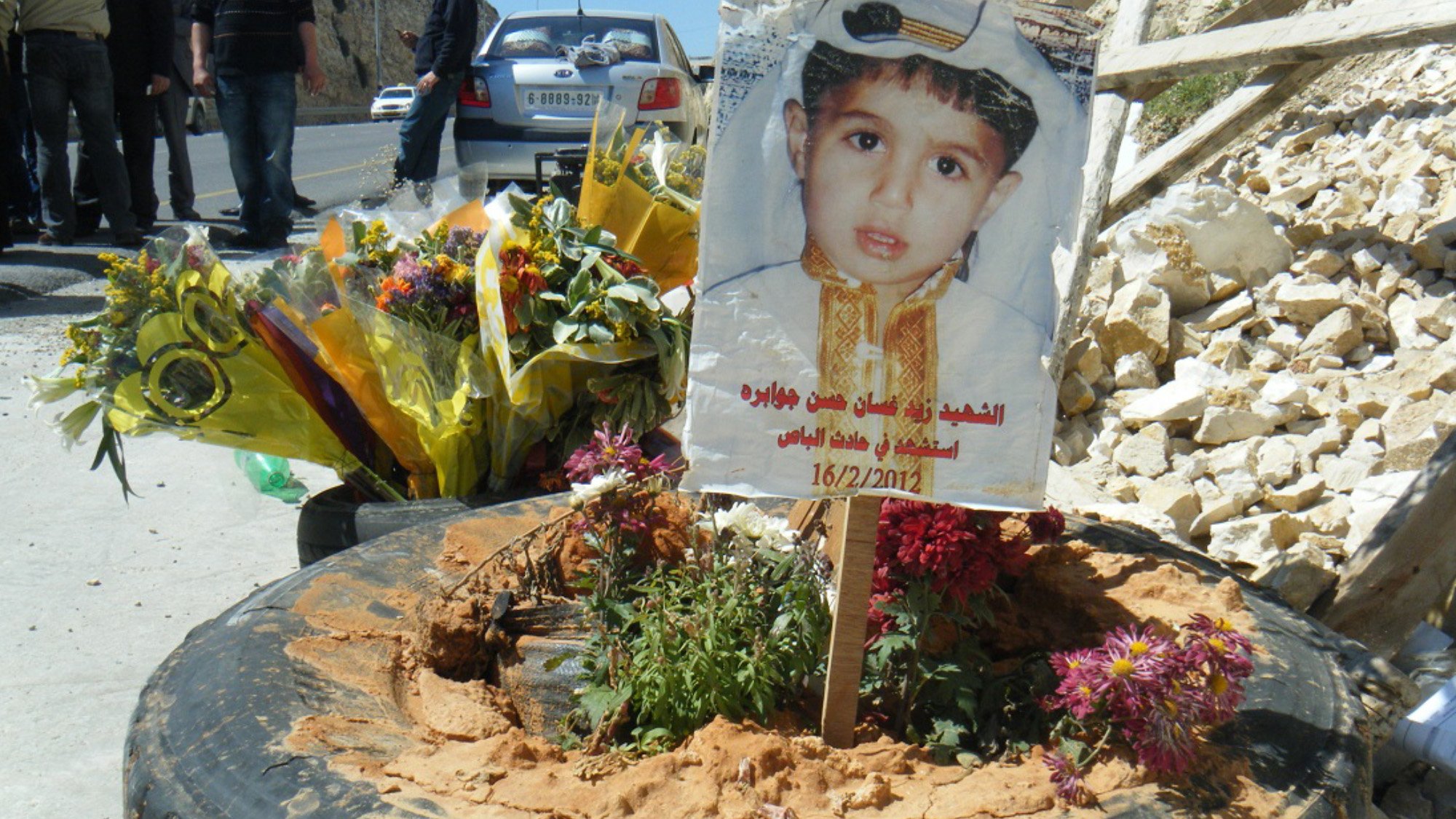 L'immagine mostra un tributo commemorativo collocato su un terreno. Al centro, c'è un cartello che presenta un ritratto di un bambino, adornato con un abbigliamento tradizionale. Attorno al cartello ci sono fiori freschi e piante, posizionati in una gomma di automobile. Il tutto è circondato da un ambiente duro, con rocce visibili e alcune persone in fondo che sembrano partecipare a una commemorazione. La data sul cartello indica un giorno significativo, possivelmente legato alla memoria del bambino.
