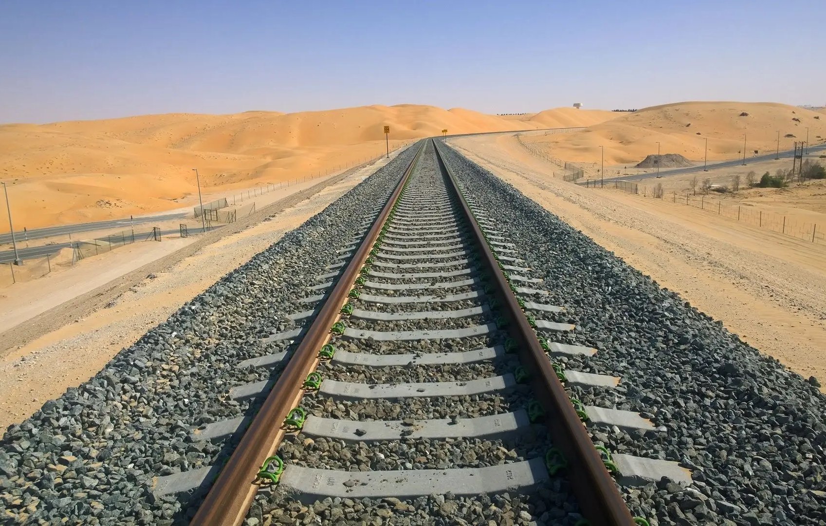 L'image montre une voie de chemin de fer s'étendant à travers un paysage désertique. Les rails, posés sur des traverses en bois, sont entourés de gravier. En arrière-plan, on aperçoit des dunes de sable sous un ciel clair et ensoleillé. Le terrain est principalement aride, et les couleurs dominantes sont des nuances de beige et de gris, ce qui souligne l'isolement et l'immensité de cet environnement désertique.