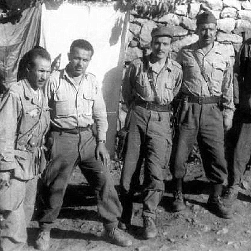 L'image montre un groupe de cinq hommes, probablement des soldats, posant ensemble devant une structure en pierre. Ils portent des uniformes militaires, composés de chemises à manches longues et de pantalons. Les expressions de leurs visages sont sérieuses et déterminées. En arrière-plan, on peut voir une toile suspendue et des pierres qui forment un mur. L'ambiance semble être celle d'un camp militaire.