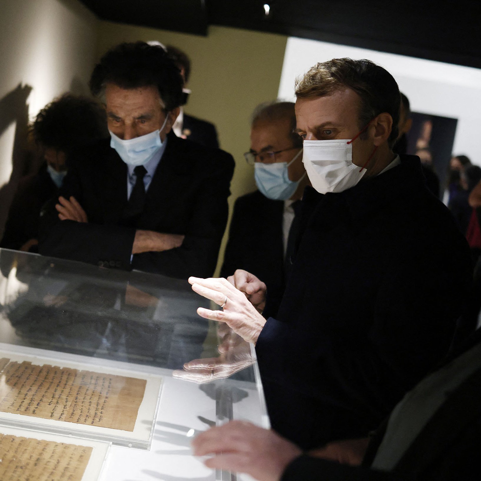 L'image montre un groupe de personnes, dont une figure centrale qui semble être un homme en train d'observer des documents exposés dans une vitrine. Ils portent tous des masques, probablement en raison de mesures sanitaires. L'environnement est sombre, avec des murs clairs, et il y a un intérêt évident pour les objets présentés. Les autres personnes dans le cadre semblent également attentives et engagées dans la discussion.