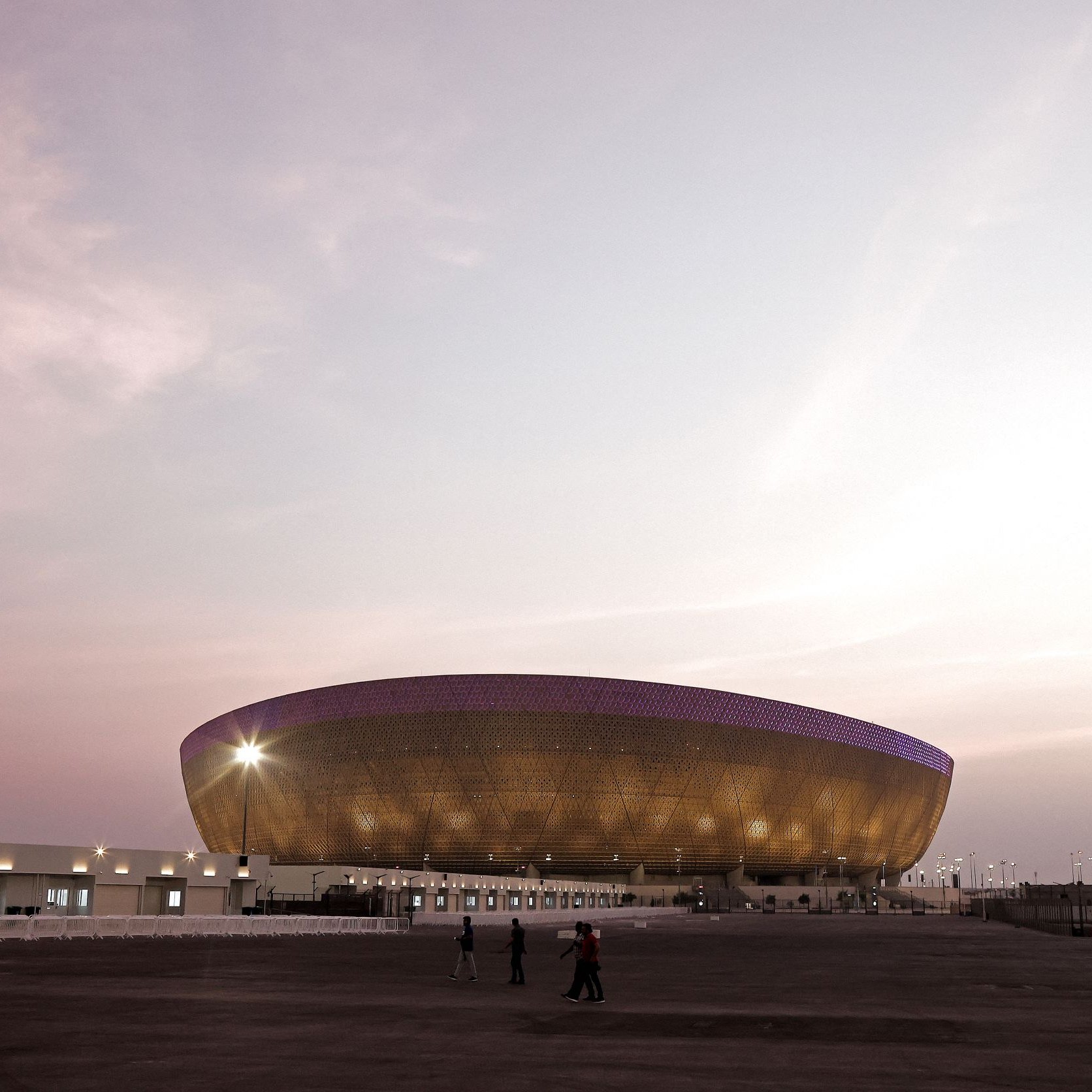 L'image montre un grand stade moderne à l'architecture distinctive, situé sous un ciel teinté de nuances pastel au coucher du soleil. Le stade possède une structure en forme d'ellipse avec une façade brillante, peut-être en métal ou en verre, qui capte la lumière ambiante. On aperçoit quelques personnes marchant devant le stade, soulignant sa taille imposante. L'ensemble de la scène dégage une ambiance calme et majestueuse.