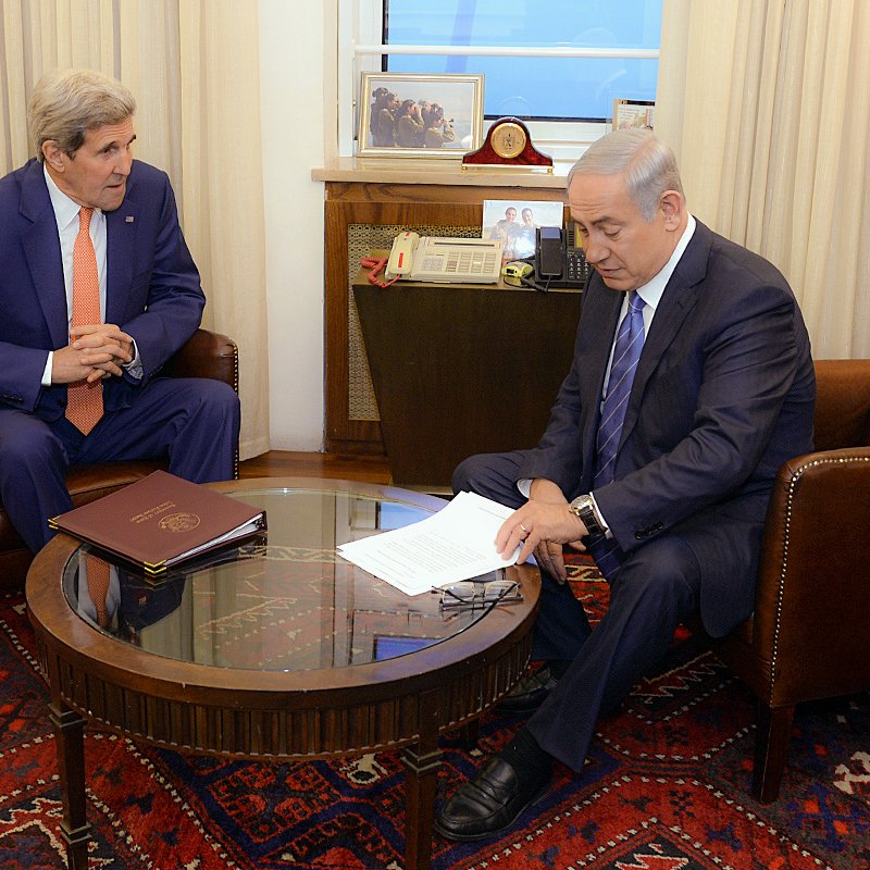 L'image montre deux hommes assis dans une pièce restructurée. L'un est vêtu d'un costume orange et l'autre d'un costume sombre, tous deux semblent être en discussion sérieuse. Ils sont assis autour d'une table basse en bois, avec des documents devant eux. En arrière-plan, on aperçoit une fenêtre et une décoration sobre, suggérant un cadre officiel et professionnel.