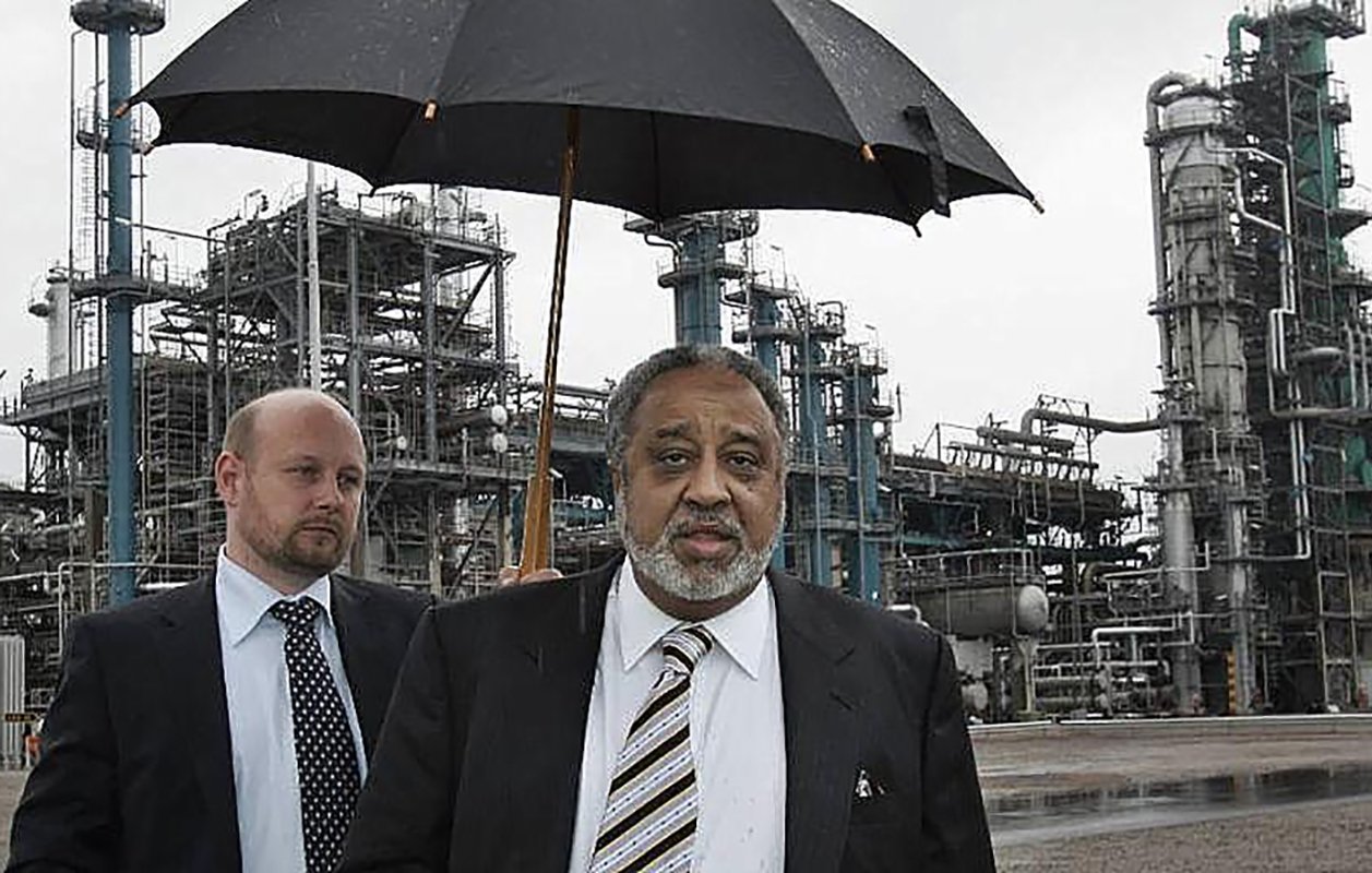 L'image montre deux hommes marchant dans une zone industrielle. L'un des hommes, qui porte un costume noir et une cravate, tient un parapluie, probablement pour se protéger de la pluie. L'autre homme, à l'arrière-plan, est également en costume. On peut voir des structures industrielles, comme des réservoirs et des cheminées, en arrière-plan, sous un ciel grisâtre. L'ambiance générale de l'image est celle d'une visite ou d'une inspection dans un environnement industriel.