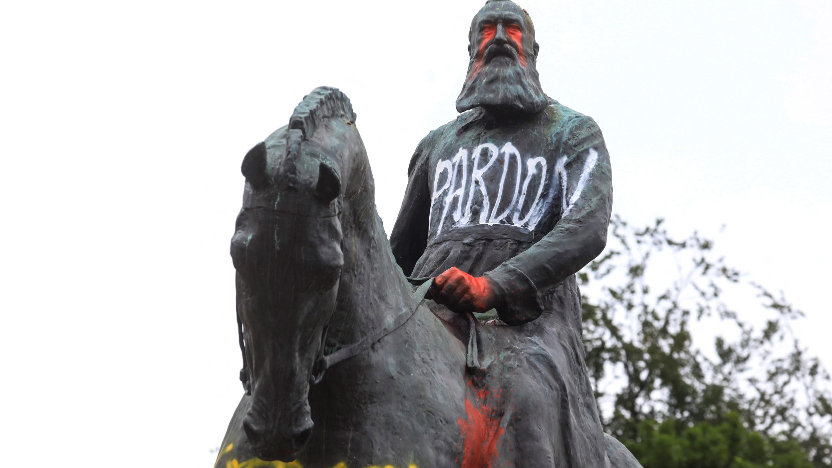 L'image montre une statue équestre d'un homme, probablement un personnage historique, qui porte des graffitis. Le mot "PARDON" est écrit en lettres blanches sur sa robe, tandis que d'autres mots, comme "RACIST", apparaissent en couleur sur le socle. La statue semble être recouverte de peinture, ce qui suggère un acte de protestation ou de critique. En arrière-plan, on peut voir un ciel nuageux et quelques arbres.