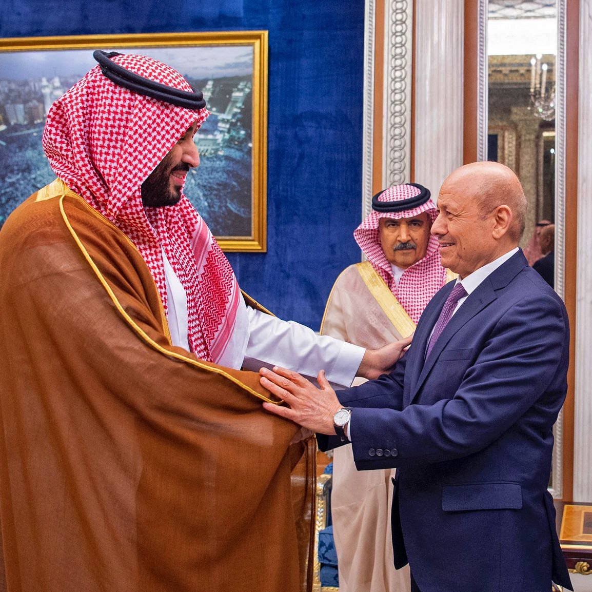 L'image montre deux hommes en costume, engagés dans une interaction amicale, probablement lors d'une rencontre officielle. L'un des hommes porte un habit traditionnel avec un châle. L'arrière-plan présente un décor élégant, typique d'un palais, avec des éléments architecturaux raffinés. D'autres personnes en tenue formelle sont visibles, suggérant un contexte diplomatique ou cérémoniel.