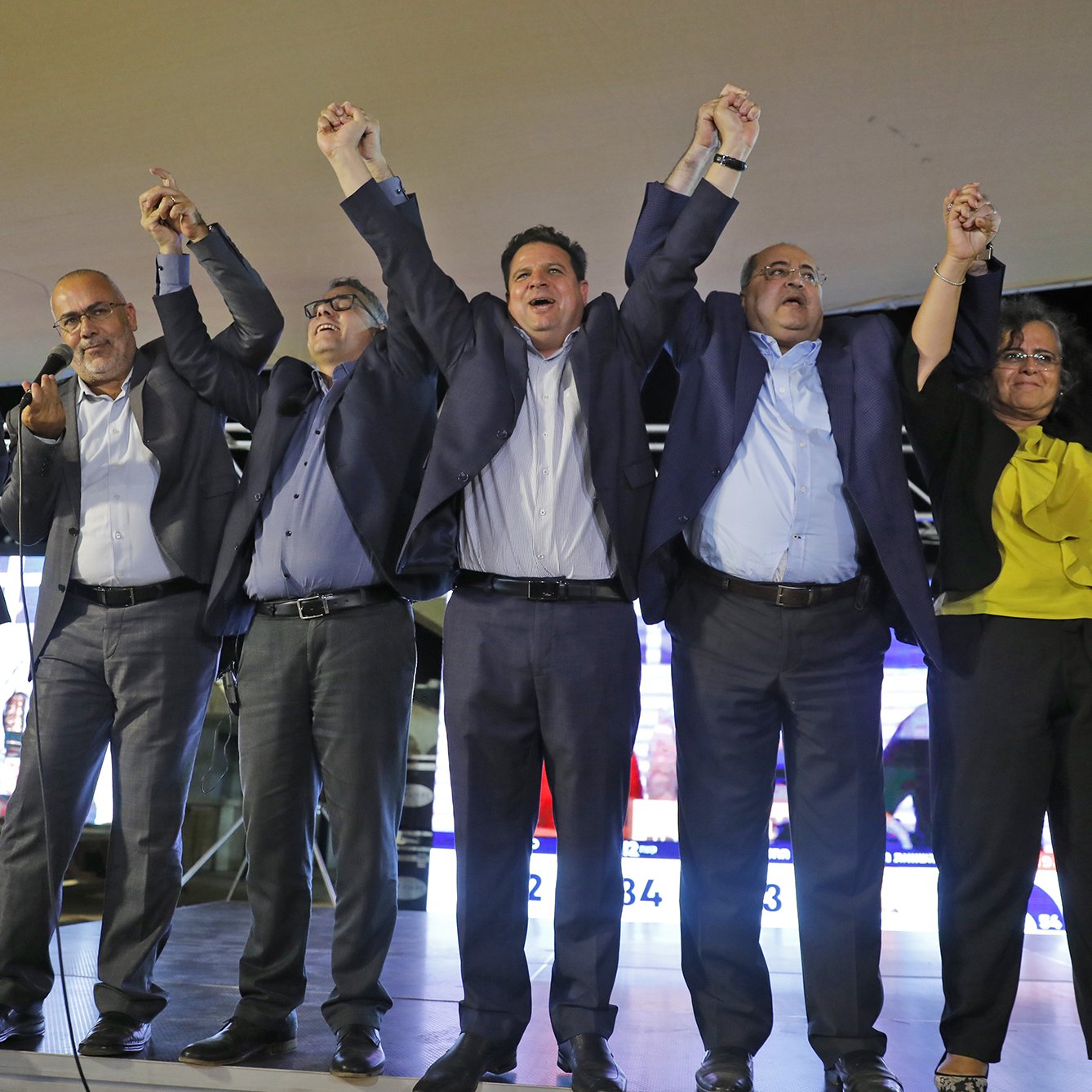 الصورة تُظهر مجموعة من الأشخاص يقفون على منصة، يرفعون أيديهم معًا في حالة من الاحتفال أو التأكيد. يبدو أنهم في مناسبة سياسية أو تجمع جماهيري، حيث تظهر خلفهم شاشات تعرض معلومات أو نتائج. يرتدي المشاركون ملابس رسمية، ويتمتعون بتعبيرات وجه تعكس الحماس والفرح.