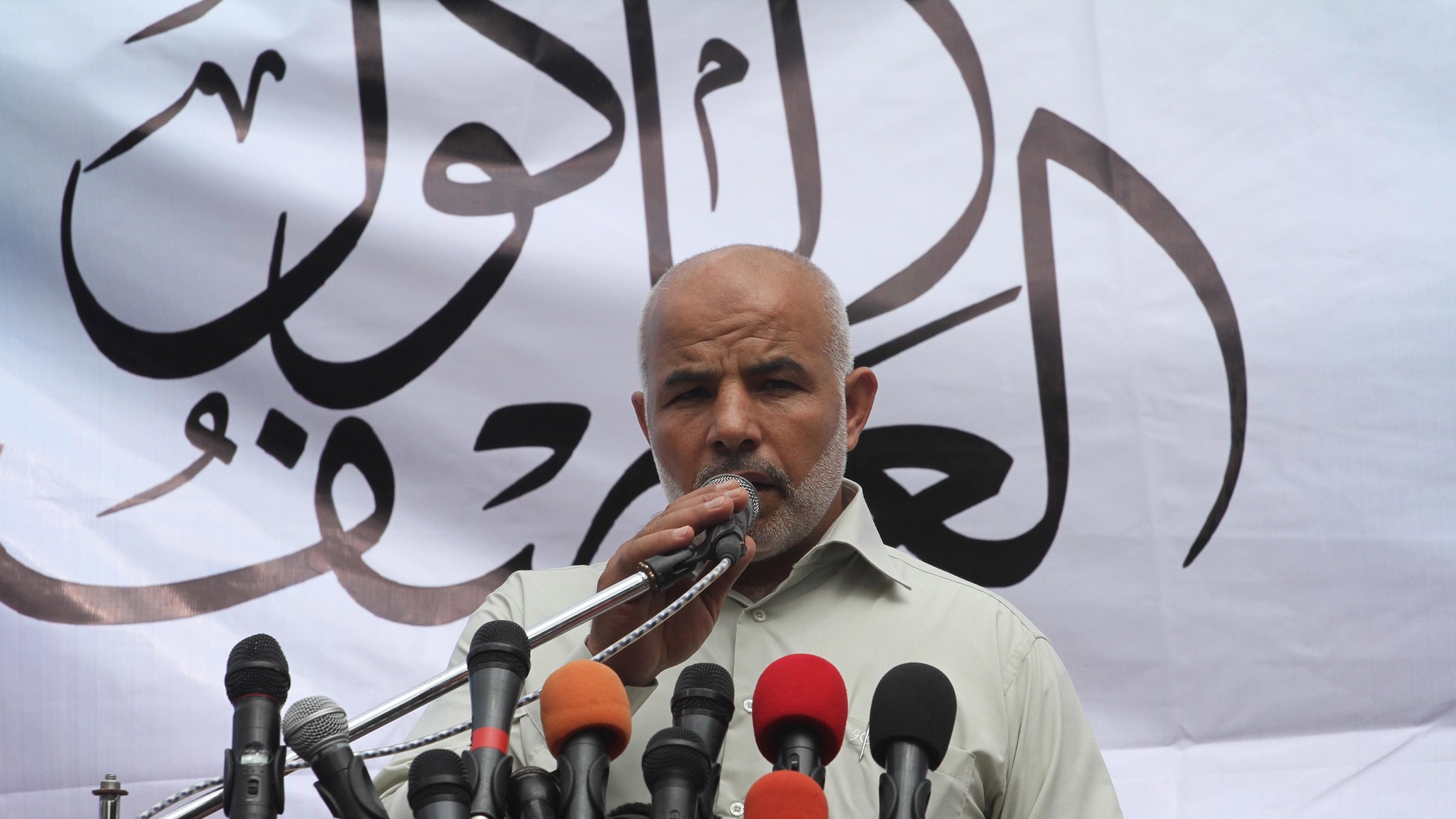 في الصورة، يظهر رجل واقفًا أمام مجموعة من الميكروفونات، حيث يبدو أنه يتحدث أو يلقي خطابًا. الخلفية تحتوي على بعض الكتابات بخط عربي، مما يشير إلى أهمية الحدث أو المناسبة. يبدو الرجل جادًا وهو يركز على توصيل رسالته للجمهور، ويعكس ذلك من خلال تعبير وجهه ولغة جسده.