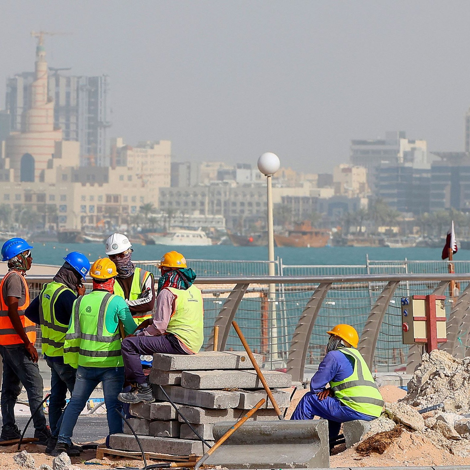 L'image montre un groupe de travailleurs sur un chantier de construction près de la mer. Ils portent des vêtements de sécurité, comme des gilets réfléchissants et des casques. En arrière-plan, on peut voir une ville avec des bâtiments modernes et des structures élevées. L'ambiance semble occupée, et le chantier est environné de matériaux de construction, avec des blocs empilés. La mer est visible au loin, ajoutant un cadre pittoresque à la scène.