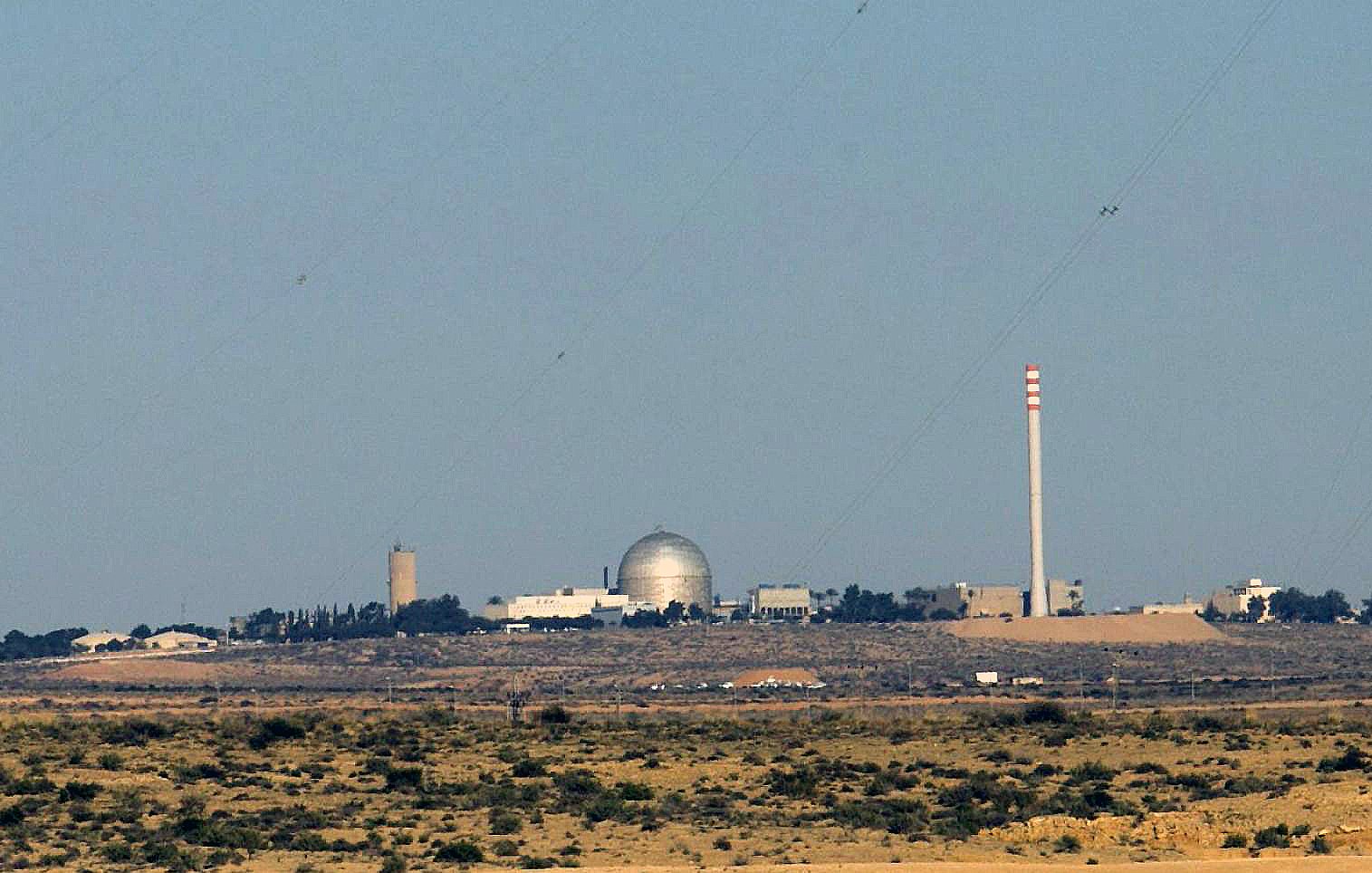 L'image montre un paysage désertique avec des bâtiments en arrière-plan. Au centre, on peut voir un dôme argenté, probablement un réacteur ou une installation scientifique. À côté, il y a une cheminée de grande taille et d'autres structures de différentes hauteurs, suggérant qu'il s'agit d'une installation industrielle ou scientifique. Le ciel est clair et il n'y a pas de nuages. Le terrain autour est aride, avec peu de végétation.
