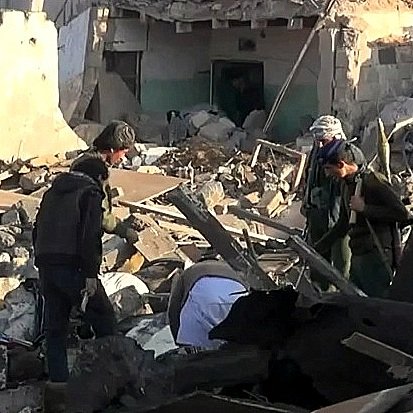 L'image montre un groupe de personnes cherchant à travers des décombres dans une zone sévèrement endommagée. Les bâtiments autour d'eux semblent en ruines, avec des murs effondrés et de nombreux débris éparpillés sur le sol. Les individus semblent concentrés et préoccupés, cherchant possiblement des survivants ou des biens perdus dans les débris. L'environnement est chaotique et témoigne d'une grande destruction.