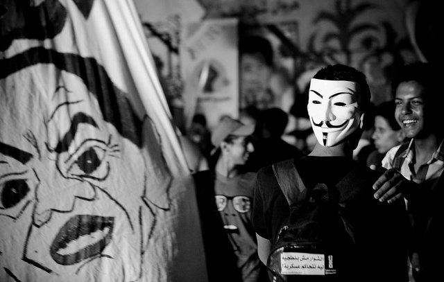 L'image montre une scène de rassemblement ou de manifestation. Un groupe de personnes est réuni, et au premier plan, une personne porte un masque stylisé, souvent associé au mouvement Anonymous. En arrière-plan, on peut voir une grande bannière avec des illustrations ou des visages, probablement liés à un message politique. L'atmosphère semble dynamique, avec des expressions de joie ou de solidarité parmi les participants. Le tout est en noir et blanc, ce qui accentue l'intensité de la scène.