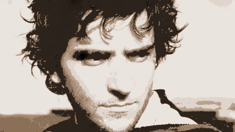 L'image montre un homme avec des cheveux ondulés et un regard intense. Son expression est sérieuse, et il semble regarder au loin, peut-être en réflexion. Le fond est flou, mettant en avant son visage. Les couleurs de l'image sont délavées, donnant une impression vintage ou artistique. Il porte un vêtement à rayures.