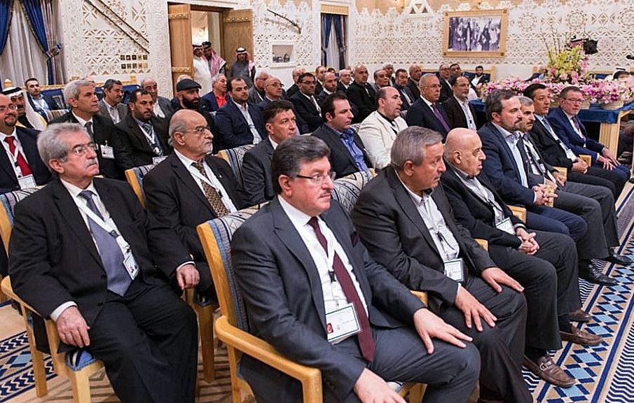 L'image montre un groupe de personnes assises, probablement lors d'une réunion ou d'une conférence. Ils semblent être des hommes d'affaires ou des responsables, vêtus de costumes formels. L'arrière-plan est orné de décorations élégantes, typiques d'une salle de conférence dans un cadre sophistiqué. Les participants regardent attentivement, suggérant un moment de discussion ou de présentation importante.