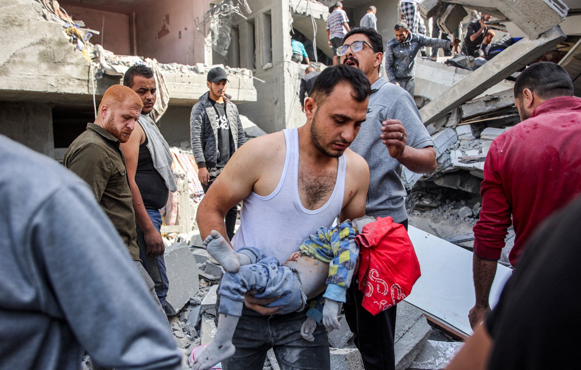 L'image montre une scène de désastre, probablement suite à un bombardement ou un tremblement de terre. On voit des décombres partout, avec des bâtiments endommagés. Un homme porte un enfant, probablement blessé, et semble préoccupé. D'autres personnes autour semblent s'affairer, cherchant à aider ou à évacuer les victimes. L'atmosphère est sombre et tragique, reflétant la détresse des personnes présentes.