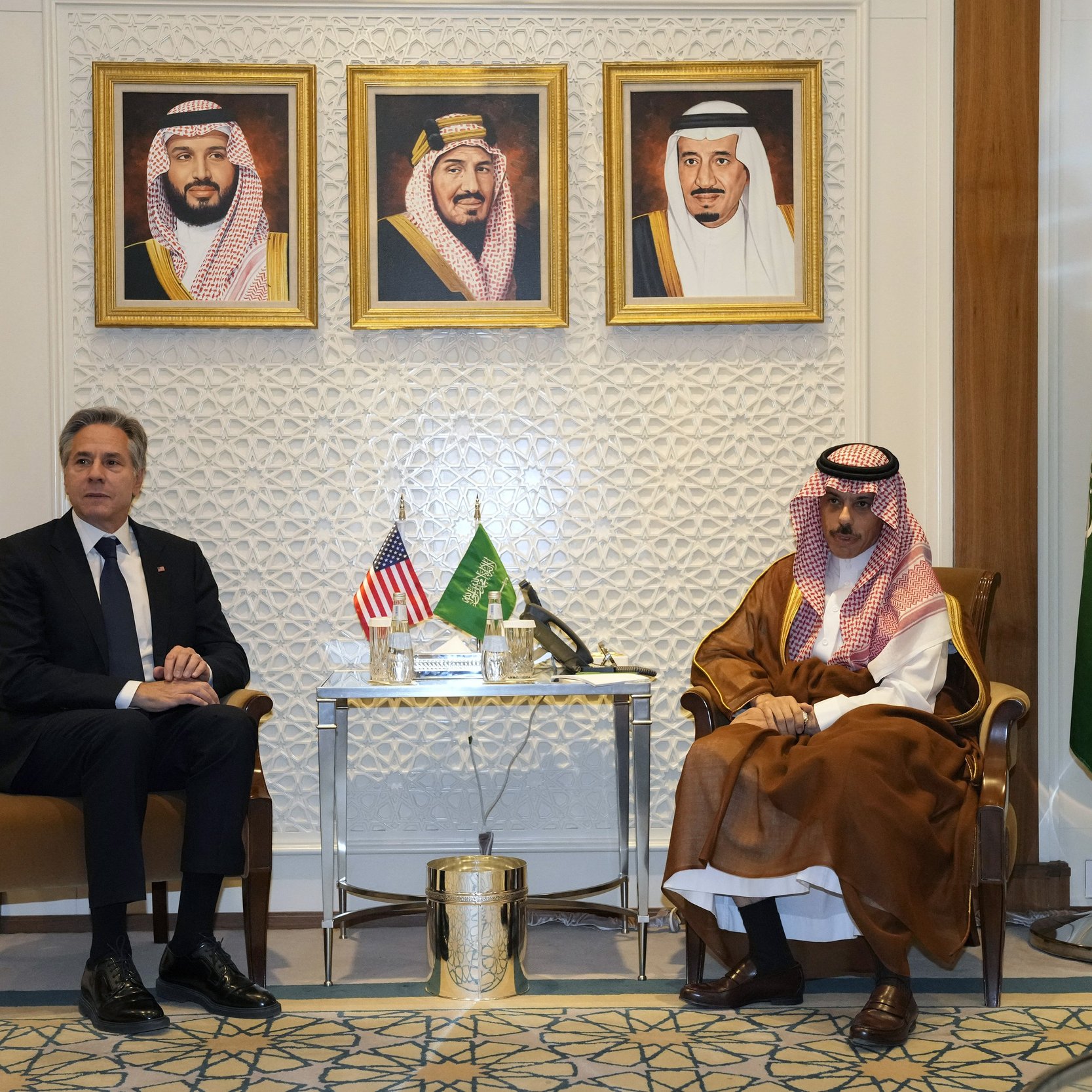 L'image montre deux hommes assis côte à côte dans un cadre formel. À l'arrière-plan, il y a trois portraits encadrés de dirigeants saoudiens. Sur une table entre eux, se trouvent des éléments décoratifs. L'un des hommes porte un costume sombre et l'autre est habillé en tenue traditionnelle saoudienne. À droite, on aperçoit un drapeau saoudien. L'atmosphère semble officielle et diplomatique.