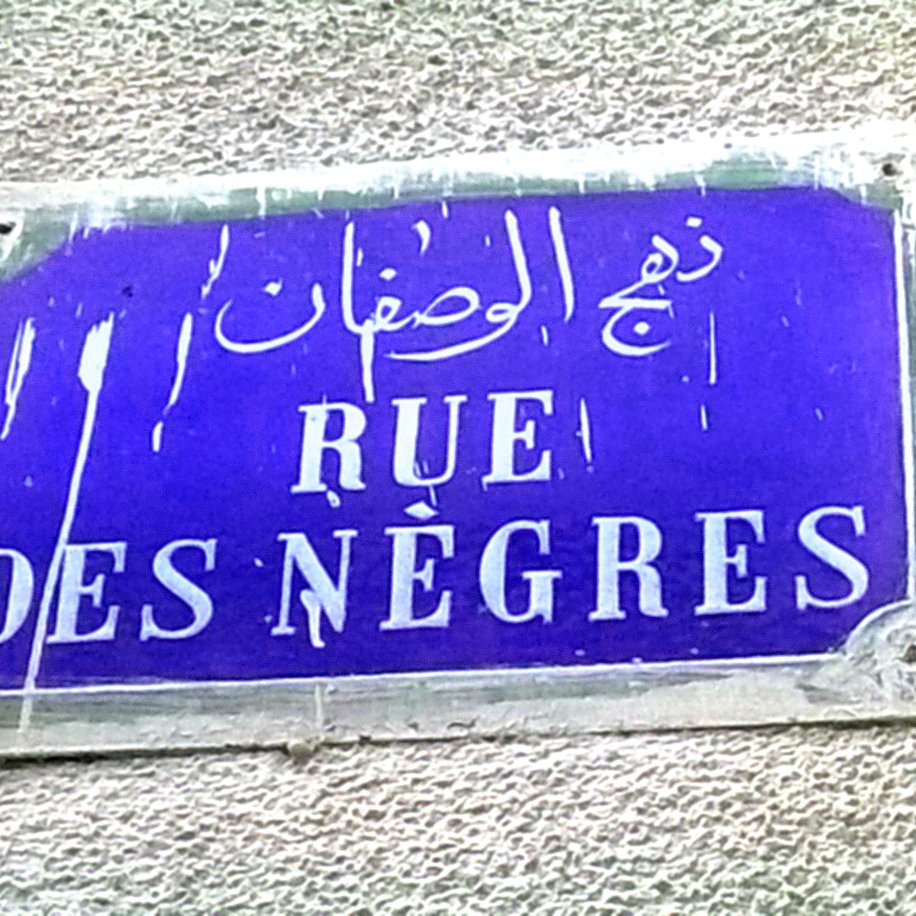 L'image montre une plaque de rue. Elle est de forme rectangulaire et de couleur bleue. Le texte inscrit est en français et en arabe. En français, il est écrit "RUE DES NÈGRES", tandis qu'en arabe, le nom de la rue est également présent. La plaque semble un peu usée, avec des marques visibles comme de l'humidité ou des éclaboussures, ce qui indique qu'elle est probablement exposée aux éléments depuis un certain temps.
