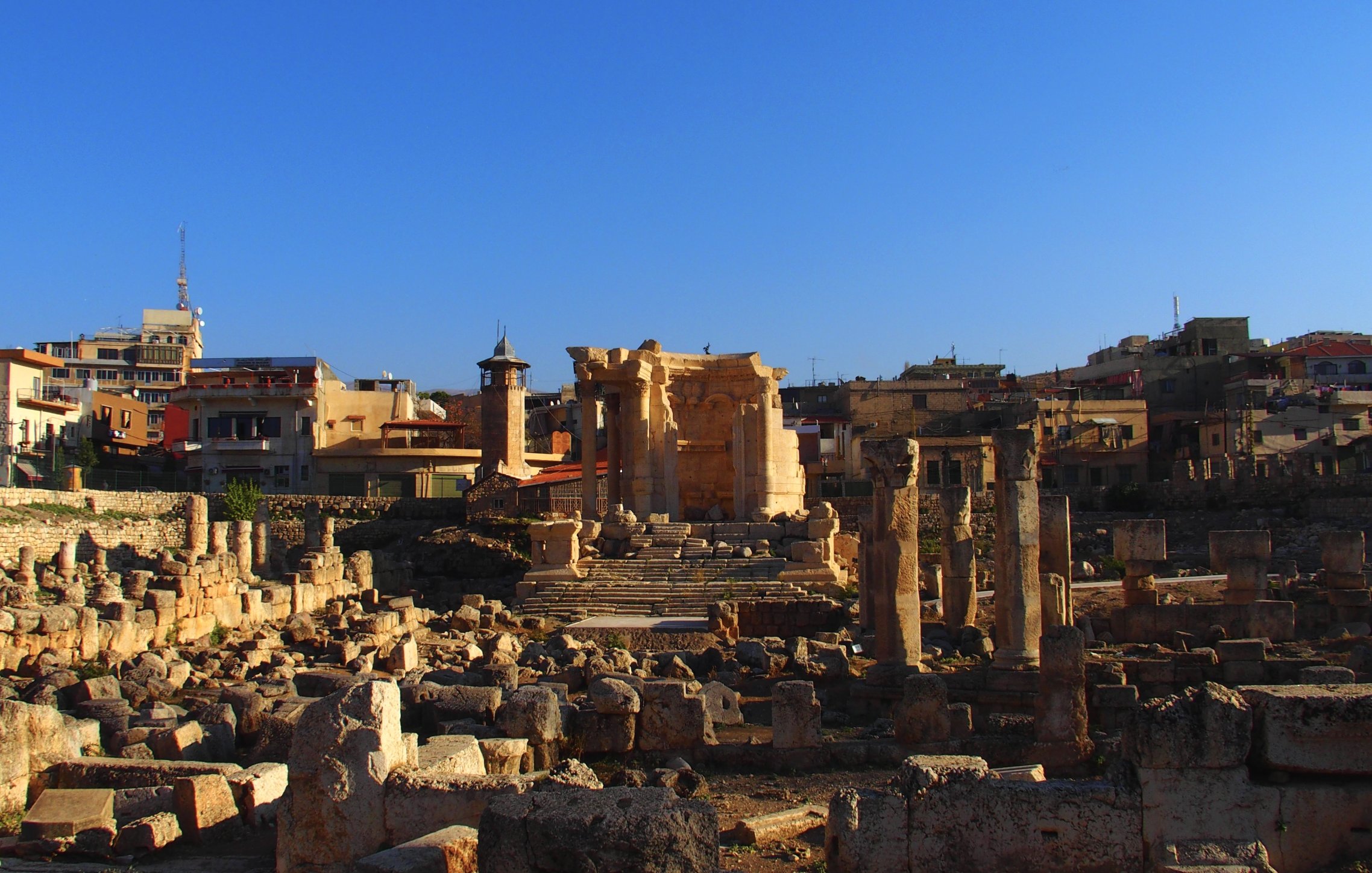 این تصویر نمایی از یک مکان باستانی را نشان می‌دهد که شامل خرابه‌های ستون‌ها و دیوارهای قدیمی است. در پس‌زمینه، چند ساختمان مدرن‌تر دیده می‌شود که نشان‌دهنده ترکیبی از تاریخ با معماری معاصر است. آسمان آبی و صاف است و نور خورشید بر روی سنگ‌ها می‌تابد که حس زیبایی و آرامش را به تصویر می‌بخشد. اجزای باستانی این مکان نشان‌دهنده تاریخ غنی و فرهنگی آنجا هستند.