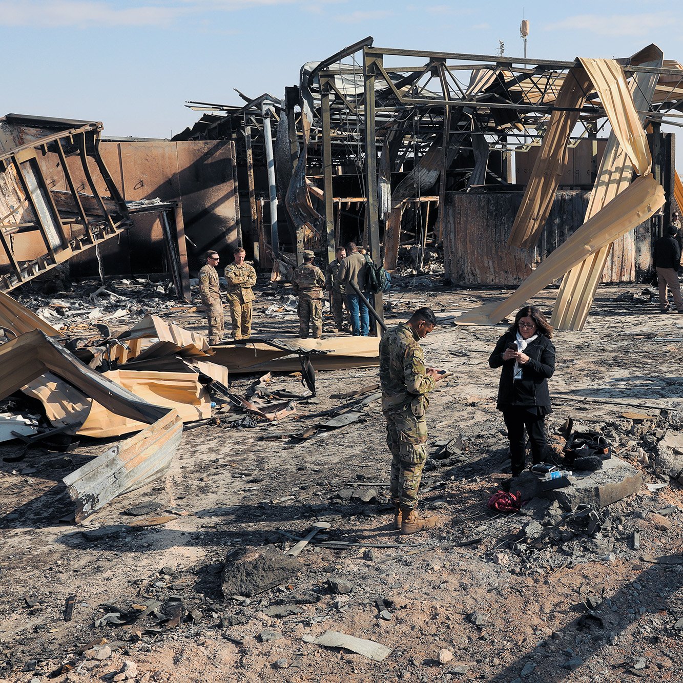 در این تصویر، صحنه‌ای از یک منطقه آسیب‌دیده به‌دلیل آتش‌سوزی یا انفجار دیده می‌شود. ساختمان‌ها به شدت خراب شده‌اند و بخشی از سازه‌ها و دیواره‌ها به شکل ناپایداری در حال افتادن هستند. چند نفر از نیروهای نظامی و خبرنگاران در میان خرابه‌ها در حال بررسی وضعیت و جمع‌آوری اطلاعات هستند. زمین پوشیده از خاک و بقایای سوخته است و آسمان آبی و صاف به نظر می‌رسد.