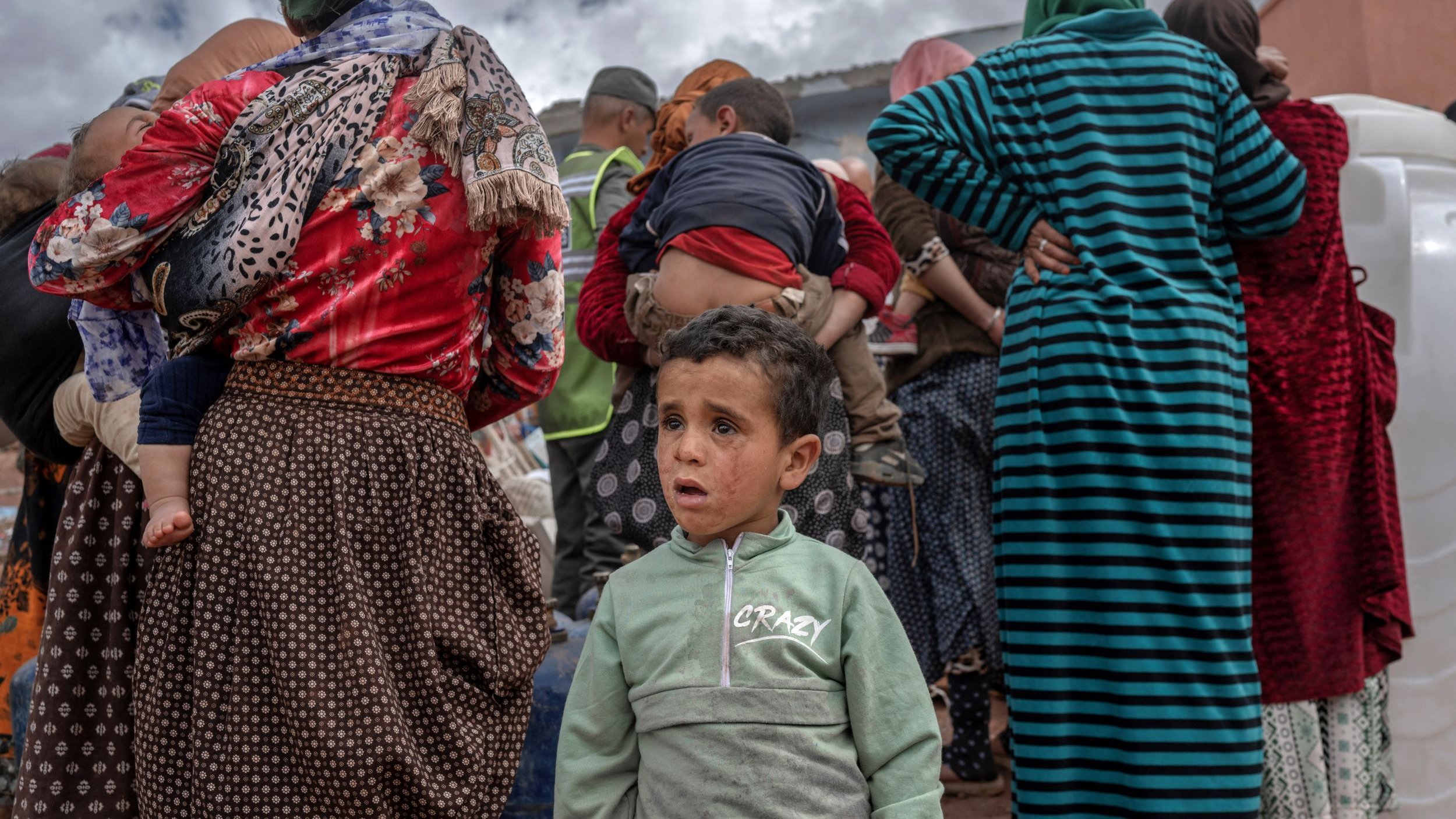 L'image montre un groupe de personnes, principalement des femmes en arrière-plan, portant des vêtements colorés et traditionnels. Parmi elles, un jeune garçon se tient au premier plan avec une expression préoccupée. Il porte une veste verte avec des écritures. Le décor suggère une atmosphère de rassemblement, peut-être dans un contexte de solidarité ou d'aide. Le ciel est nuageux, ajoutant une certaine gravité à la scène.