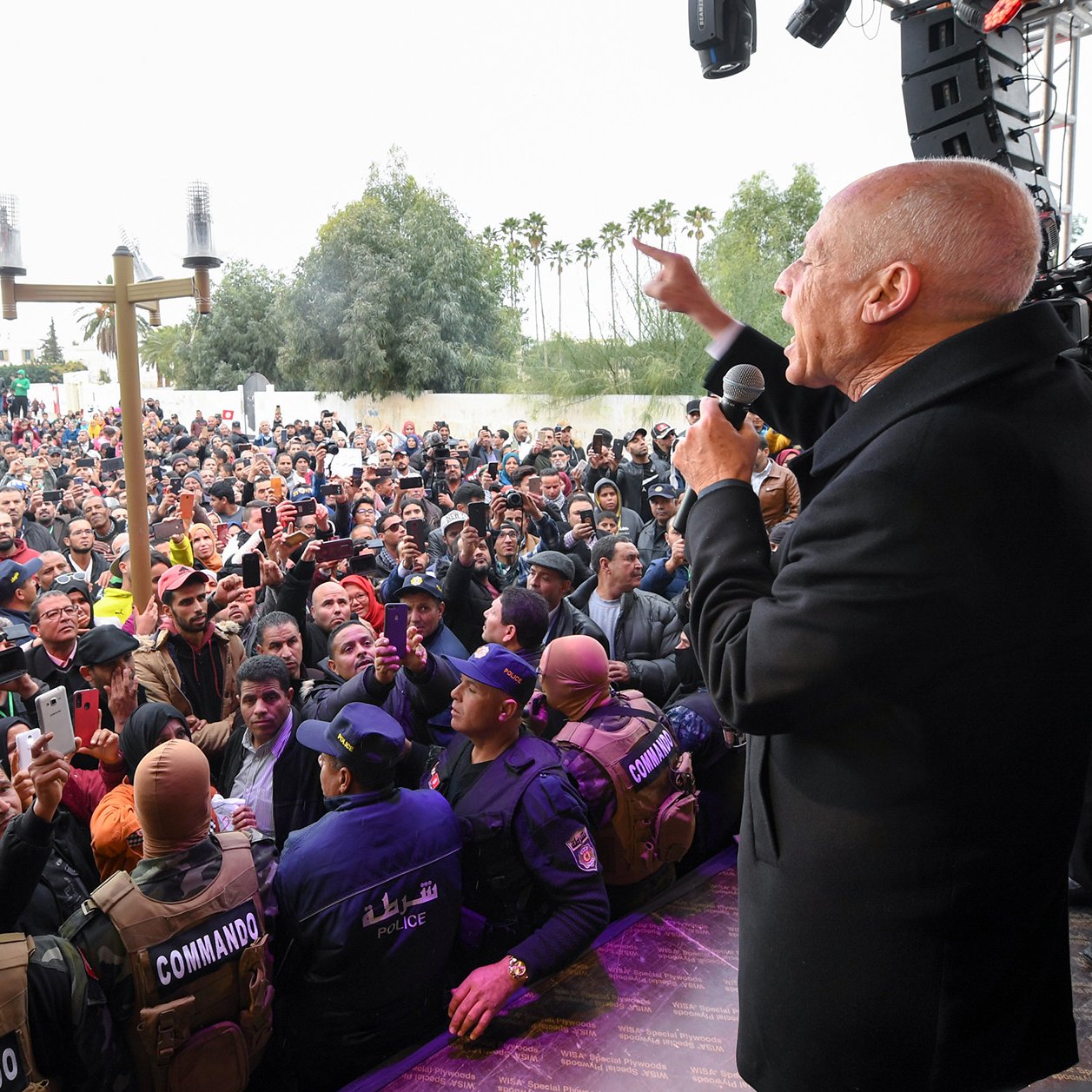 L'image montre un homme, probablement un orateur, s'adressant à une grande foule de personnes. La scène semble se dérouler à l'extérieur, avec des arbres en arrière-plan. Le public est très nombreux et semble attentif à ce qui est dit. À l'avant de la scène, des membres des forces de sécurité ou des agents en uniforme sont présents pour assurer l'ordre. L'atmosphère paraît dynamique et engagée, suggérant un événement public important.